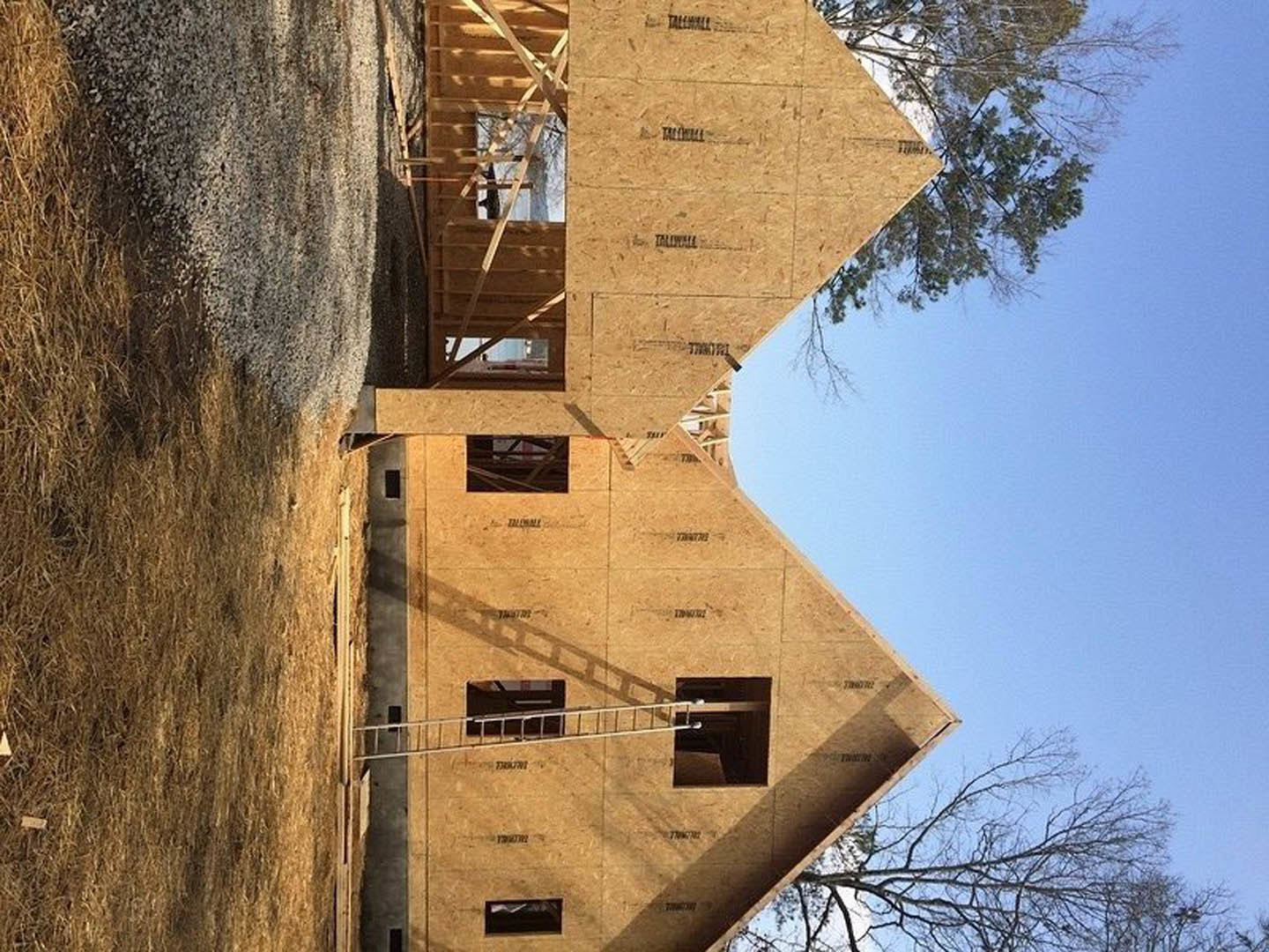 Wood-framed house under construction with exposed brick walls, large window opening, and tree in the background