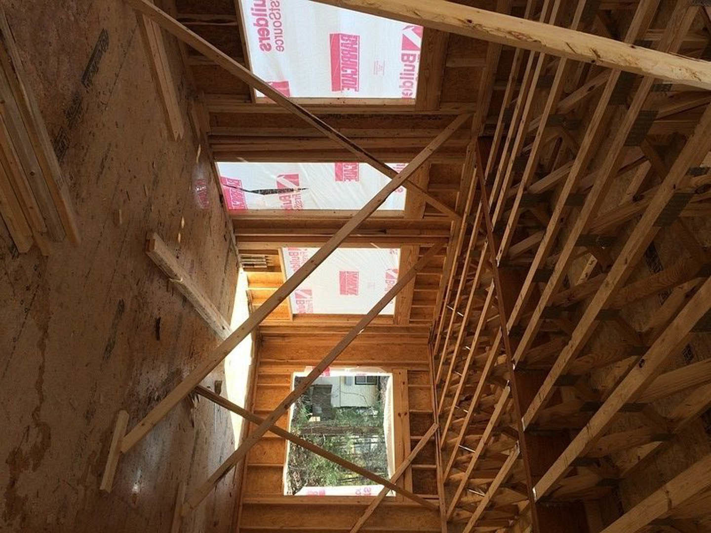 Exposed wooden framing and ceiling beams inside a house under construction, with insulation visible between studs and a window opening in the background.