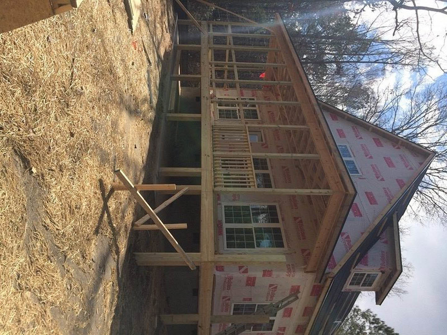Wood-framed house under construction with red and white insulation, exposed beams, multi-pane window, and front porch structure in a grassy field.