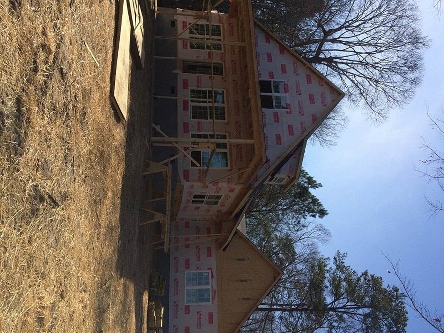Wood-framed house under construction with exposed beams, red and white safety tape, unfinished roof, and front porch; surrounded by bare trees and winter landscape.