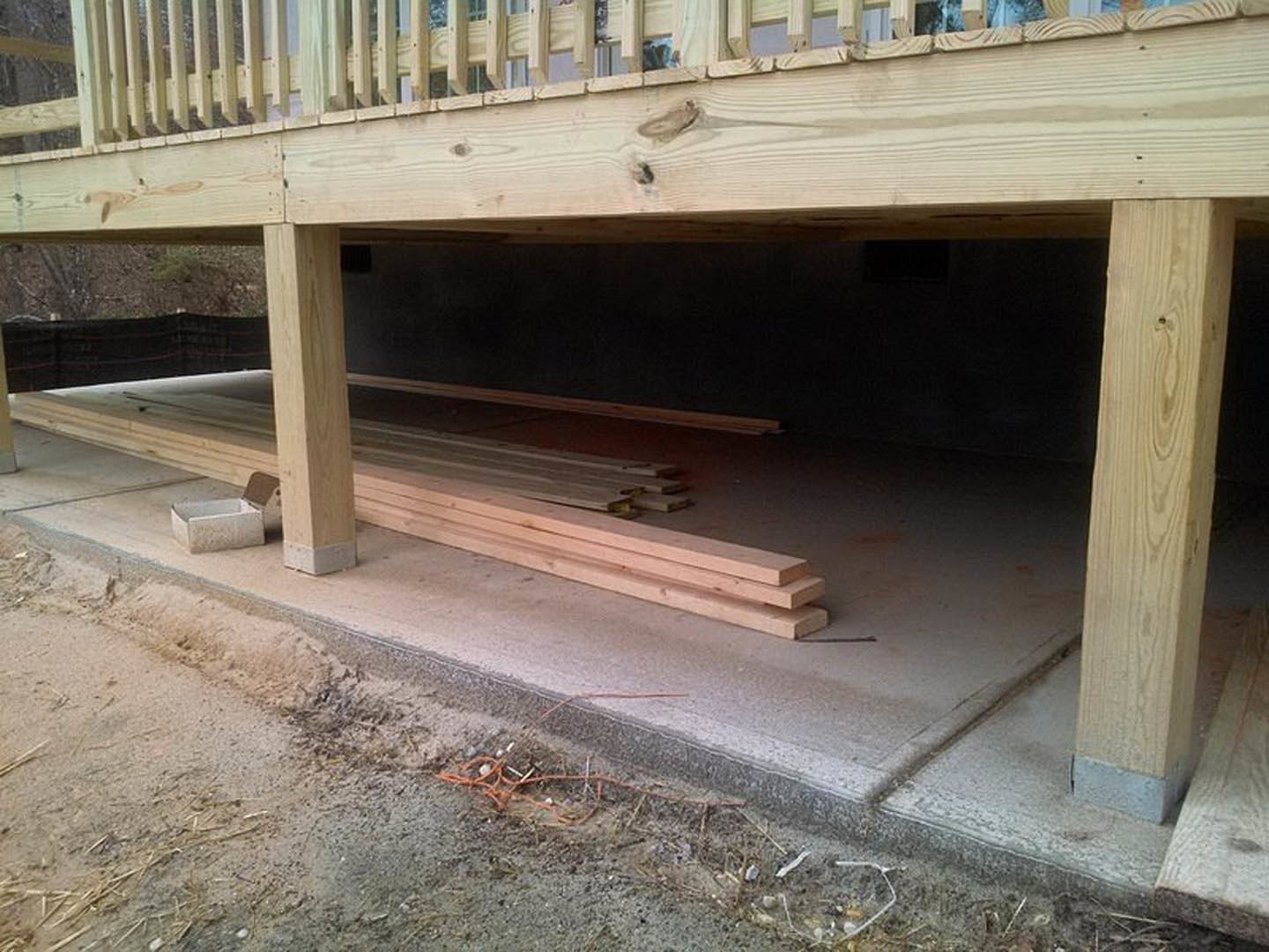 Lower deck with wood plank flooring beneath an upper deck, wood wall siding, stacked lumber on concrete ground, outdoor bench, and composite fence in background