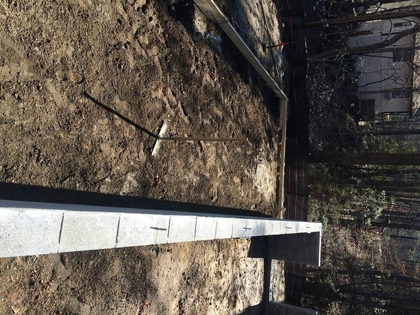 Concrete pillar set in dirt near white brick, with white house and bare trees in wooded background; blurry person walking outdoors.