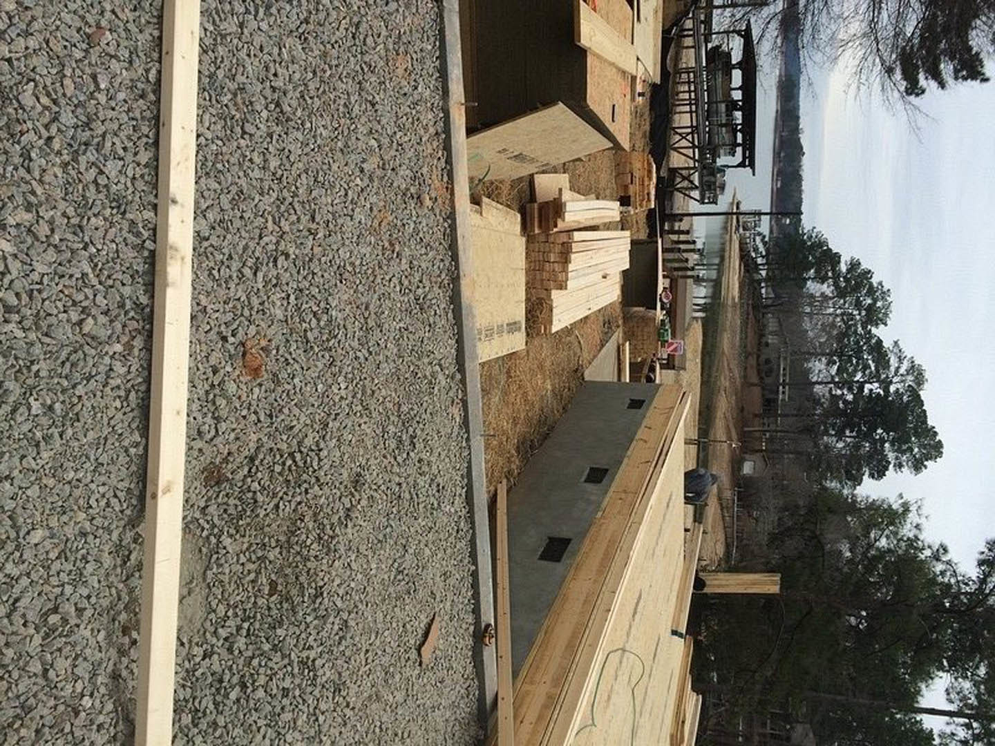 Stack of wood planks resting on gravel driveway beside unfinished concrete floor with vent holes, outdoor construction materials visible under open sky.
