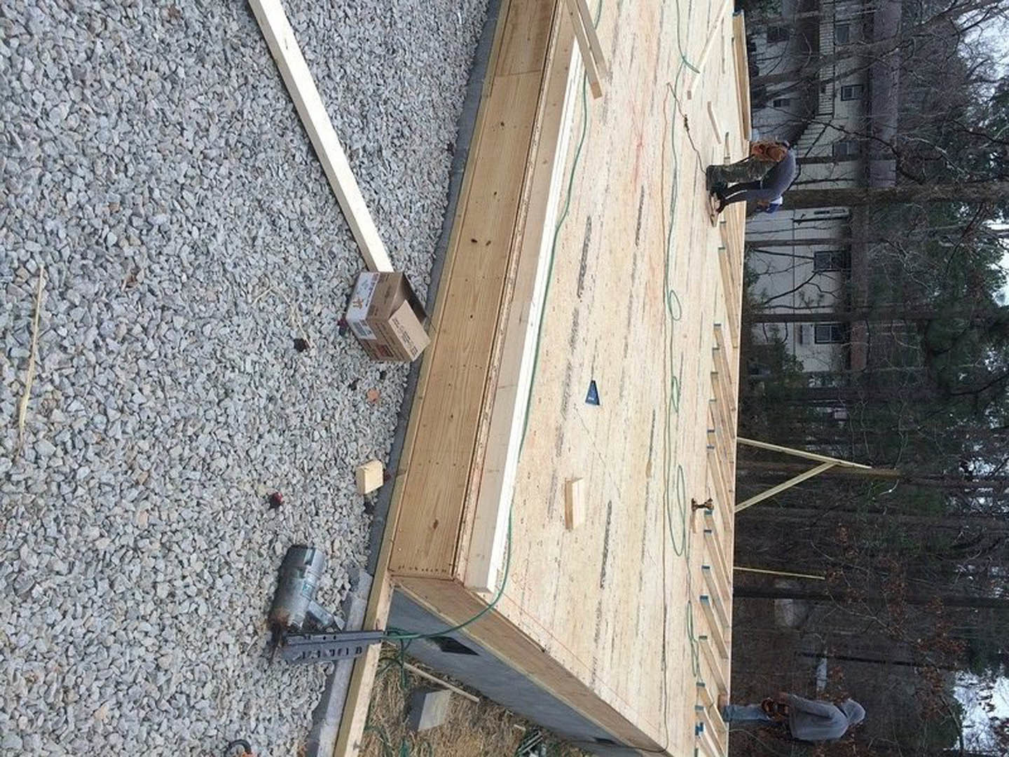 Man in military uniform standing on unfinished wooden roof with house framing visible, surrounded by trees and construction materials