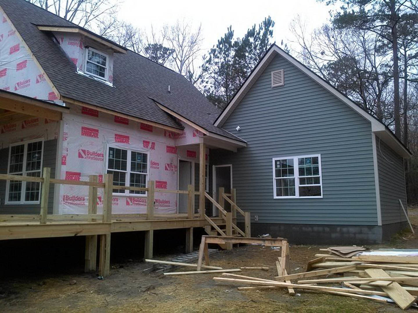 Two-story house under construction with exposed wood framing, unfinished siding, large windows, wooden porch with railing, and stacks of lumber on the ground.