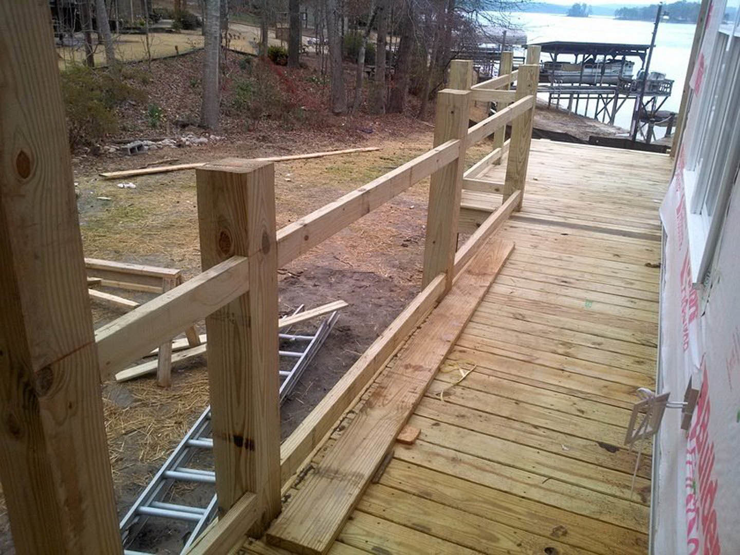 Wooden deck with horizontal railings overlooking a dock, surrounded by trees and sky, with plank flooring and lumber construction.