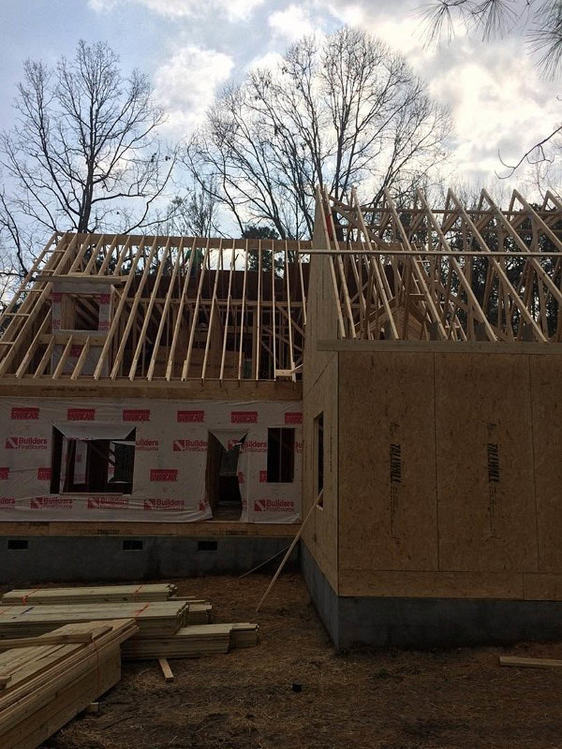 Wood-framed house under construction with exposed beams, ladder leaning against structure, leafless tree nearby, white sheet partially covering exterior, cloudy sky overhead