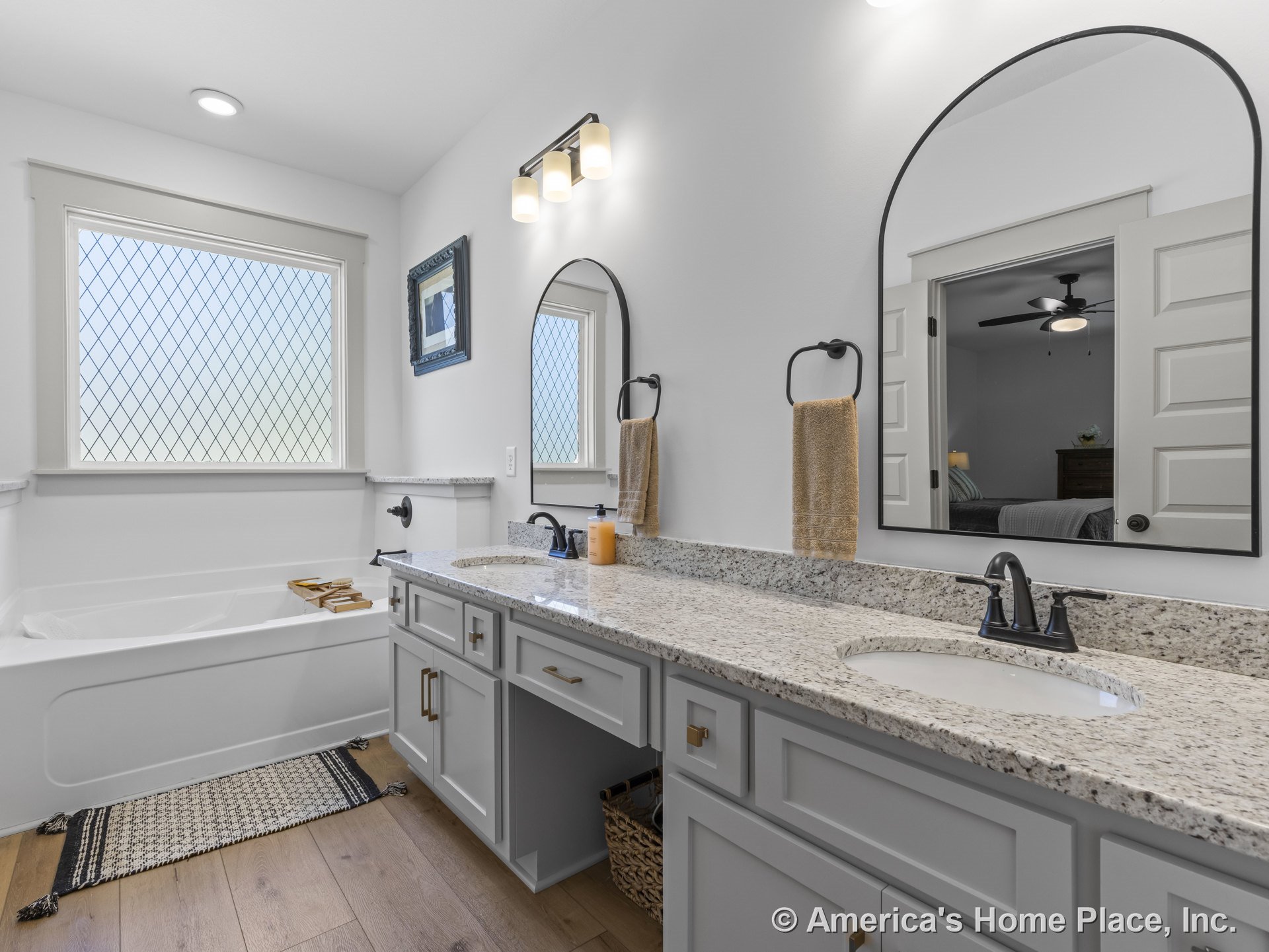 Double-sink vanity with granite countertops, built-in bathtub beneath a large privacy window with grid pattern, wood flooring, wall-mounted lighting fixtures, framed mirrors, white