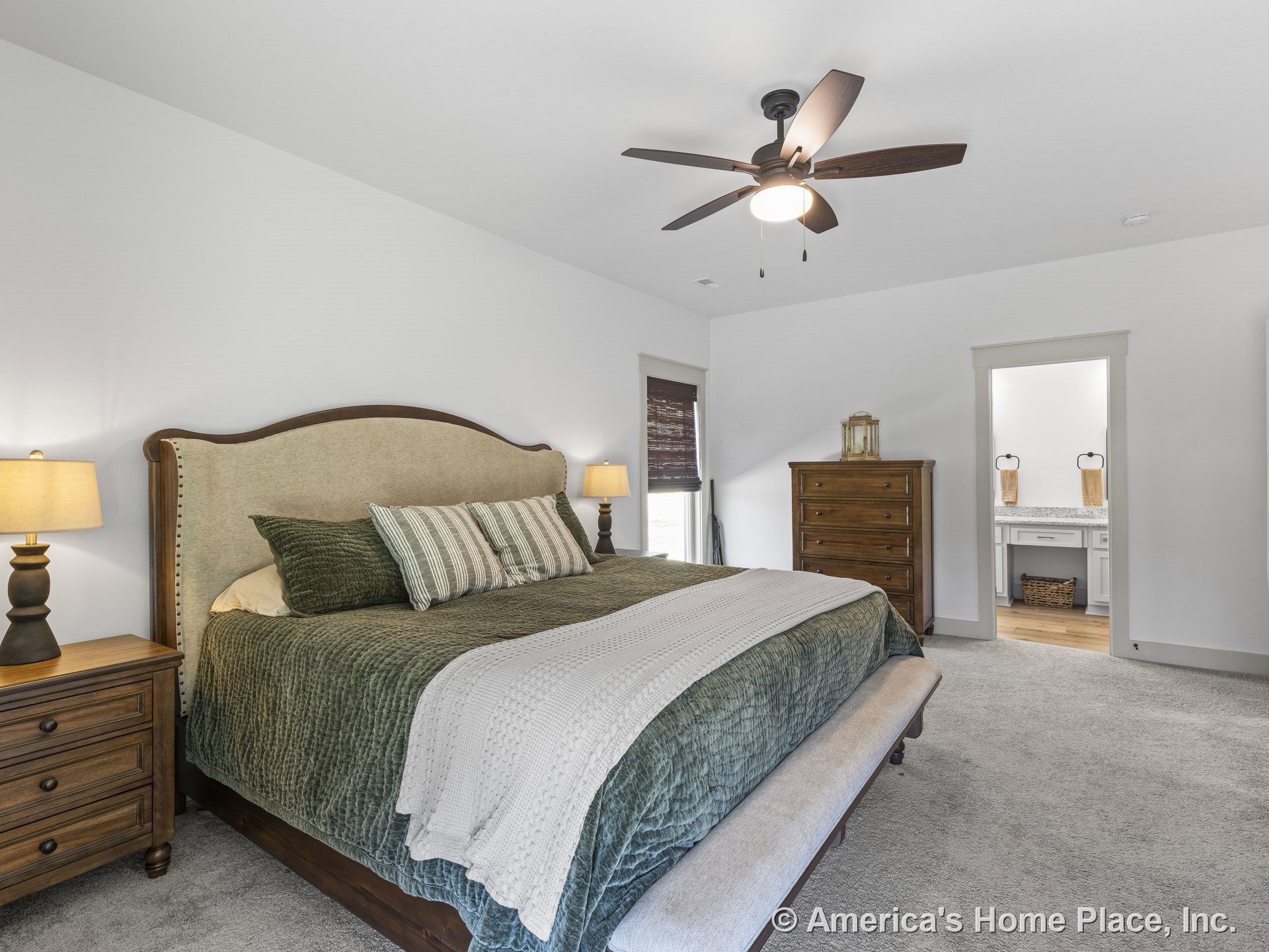 Carpeted primary bedroom with ceiling fan and built-in light, trimmed doorway leading to en suite bathroom featuring granite countertop, wood cabinetry, and white painted walls.