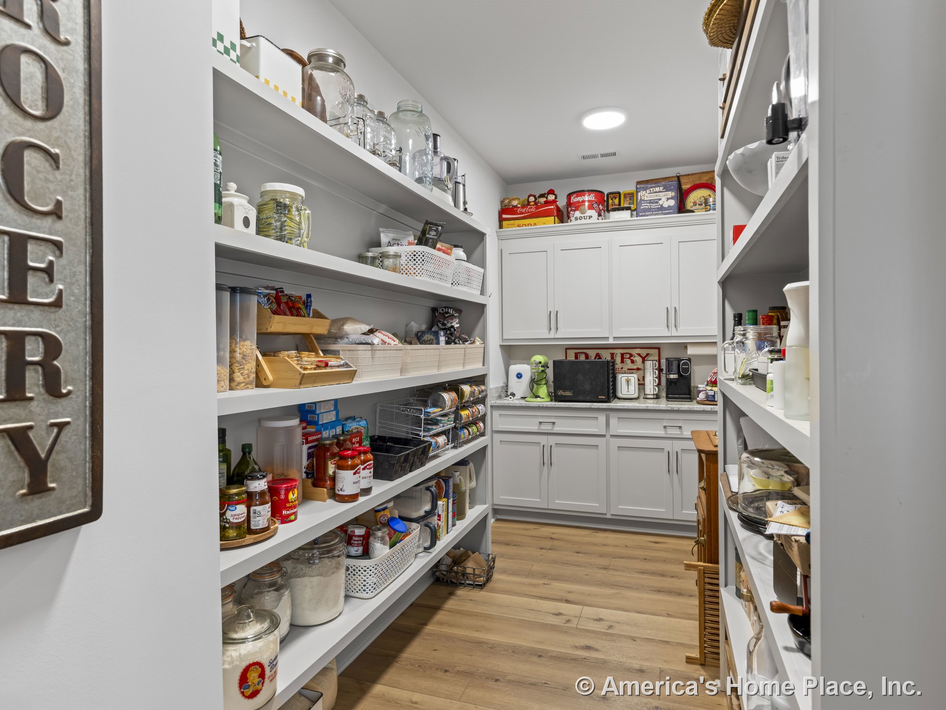 Walk-in pantry with white built-in shelving, gray shaker cabinets, wood plank flooring, recessed ceiling light, and organized storage layout with cabinet hardware.
