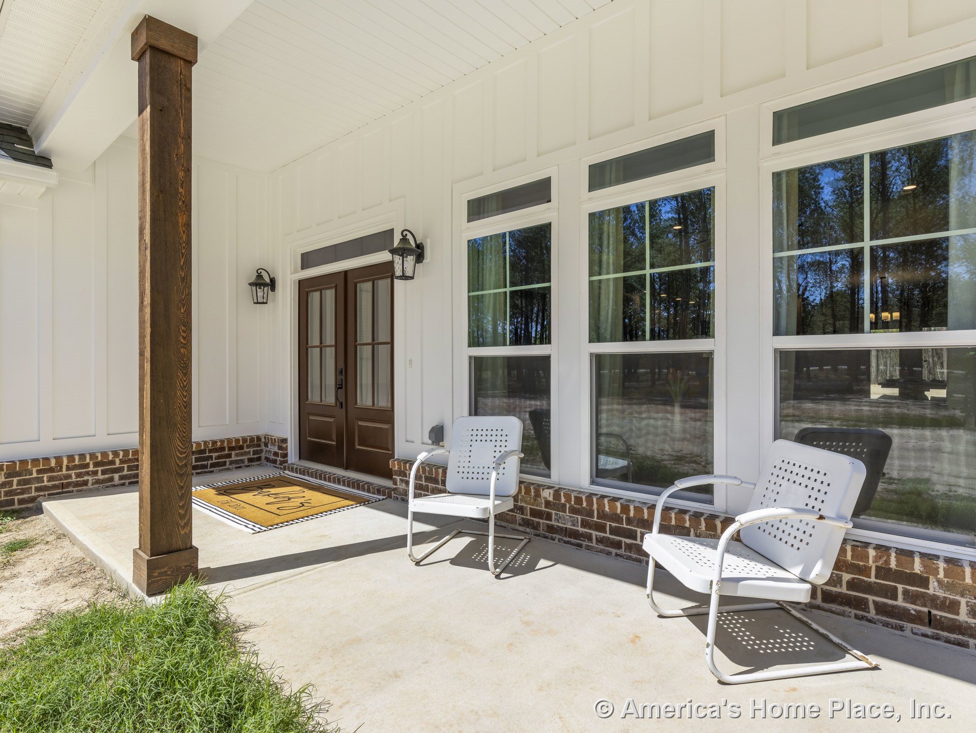 Covered front porch with wood column, brick foundation trim, double glass-panel front doors, large grid windows, white vertical siding, built-in porch lighting, transom windows