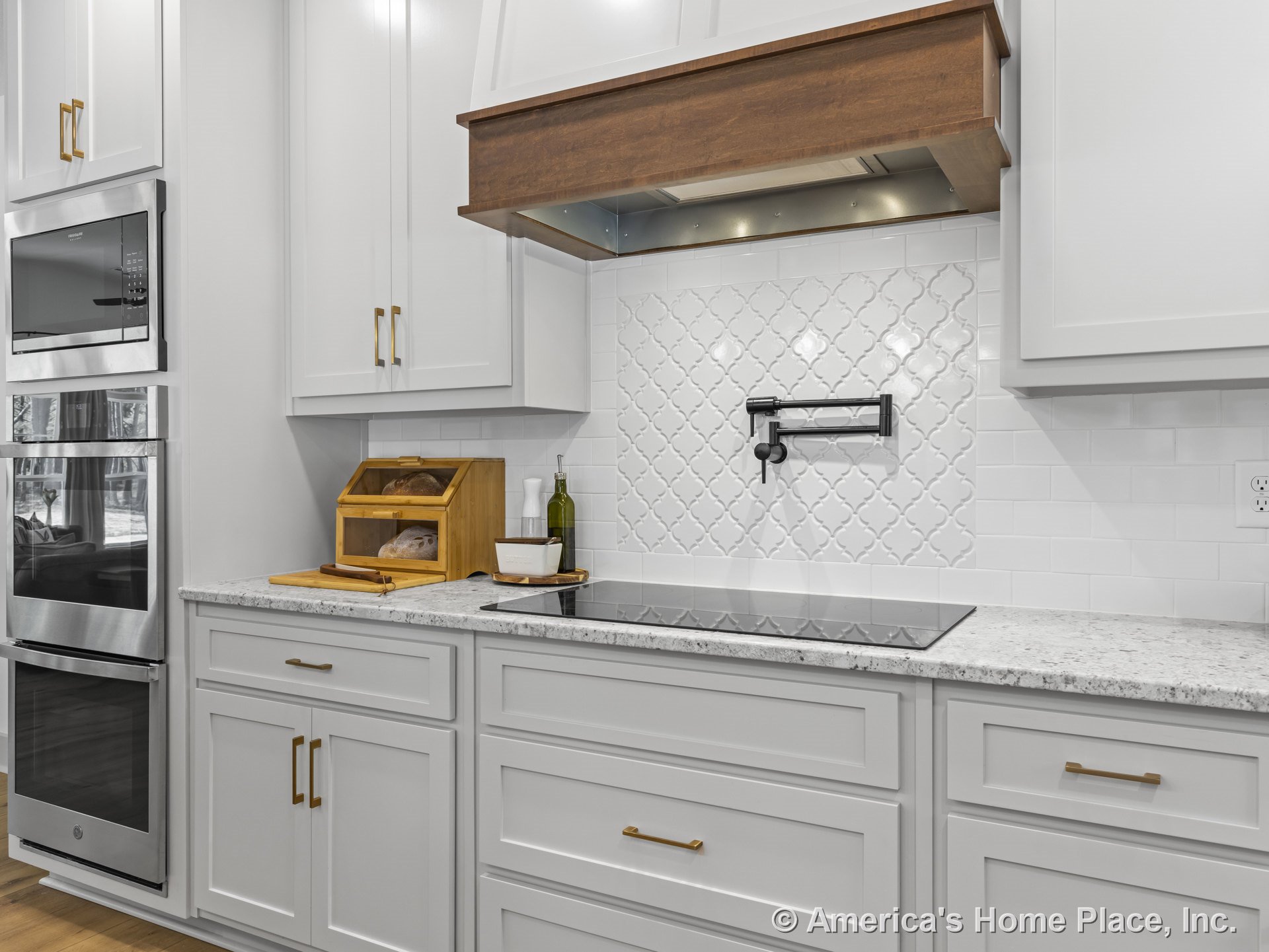 White shaker cabinets with brushed drawer pulls, patterned tile backsplash, granite countertops, built-in pot filler above the cooktop, wood range hood, and double wall ovens