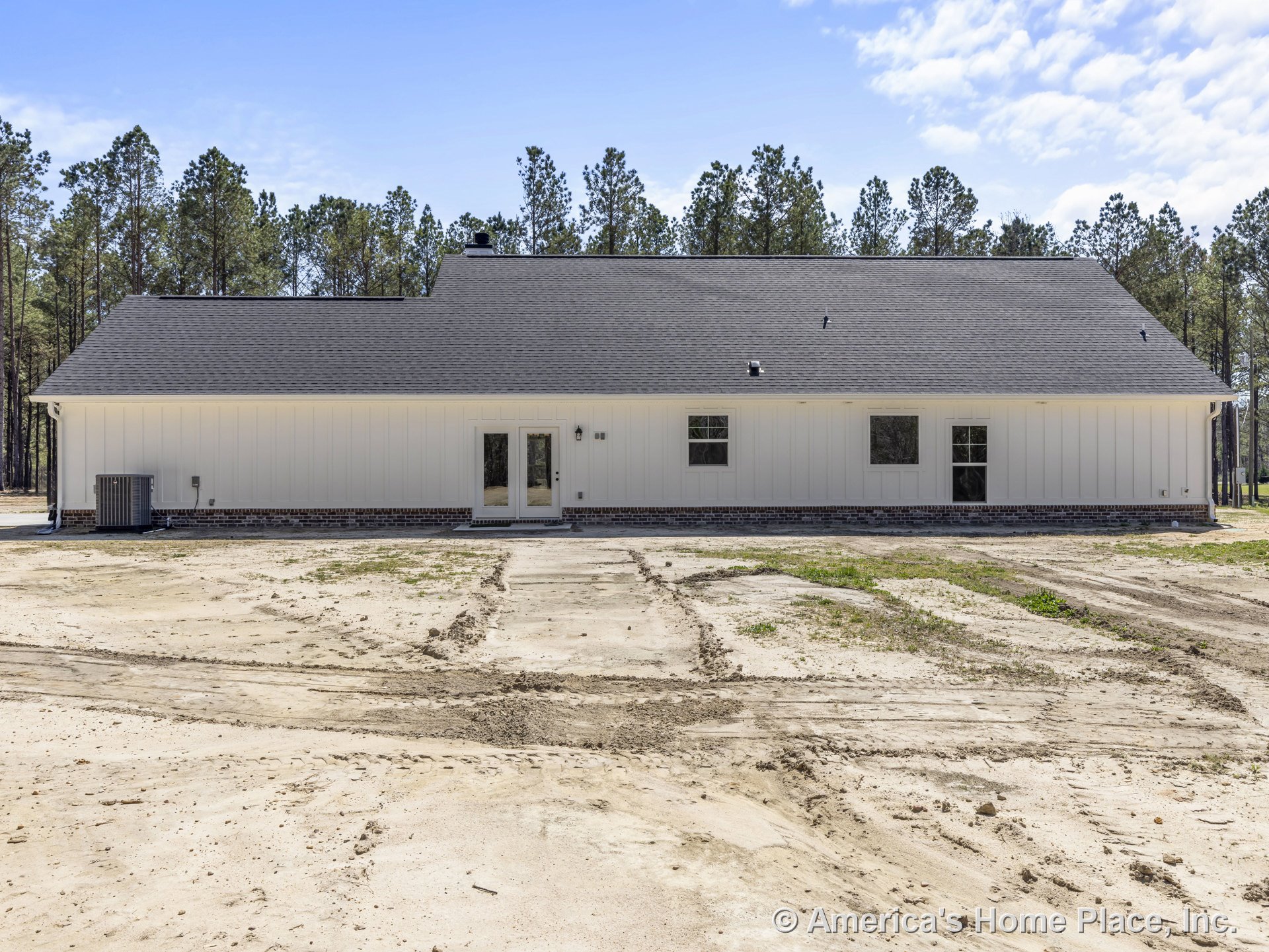 White vertical board and batten siding with brick foundation trim, dark shingle roof, double glass doors, multiple rectangular windows, and exterior wall-mounted light fixture.
