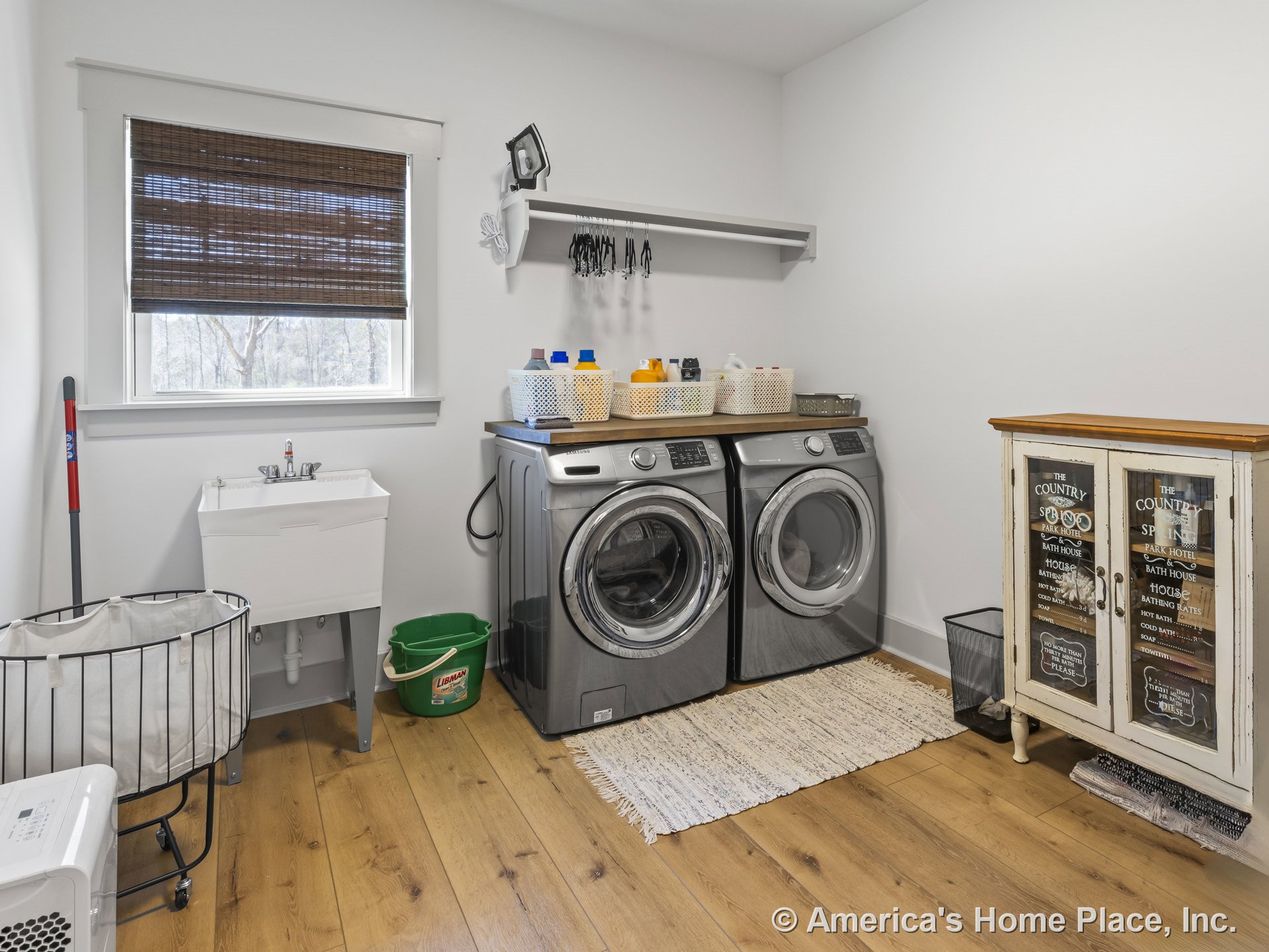 Front-loading washer and dryer beside a utility sink with chrome faucet, wood plank flooring, window with woven shade, open wall shelf, and painted cabinet with glass doors against