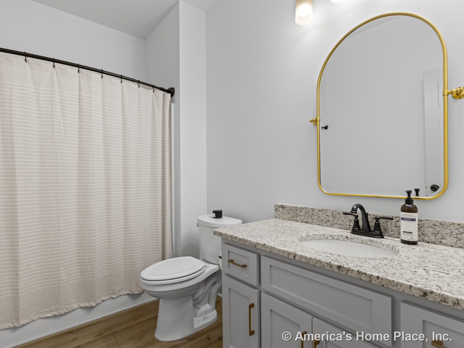 Granite vanity with light gray cabinets, curved gold-framed mirror, black faucet, wood-look flooring, wall-mounted lighting, shower curtain, and white toilet.