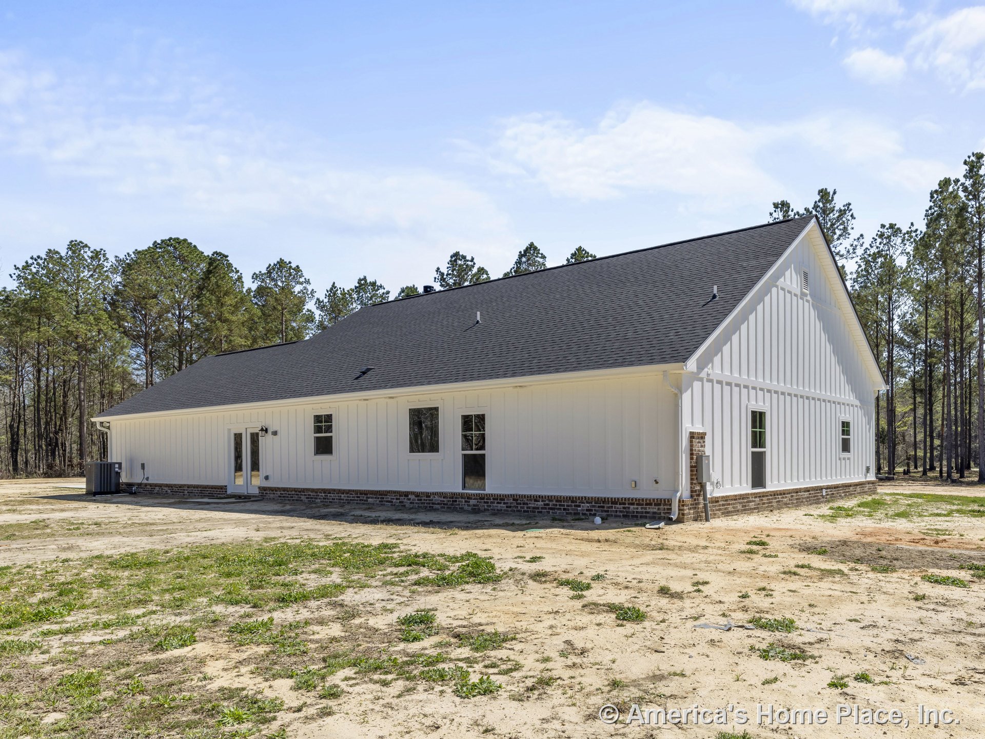 White vertical board and batten siding with brick foundation trim, black shingle roof, multiple rectangular windows, glass-paneled exterior door, and outdoor lighting fixture on a