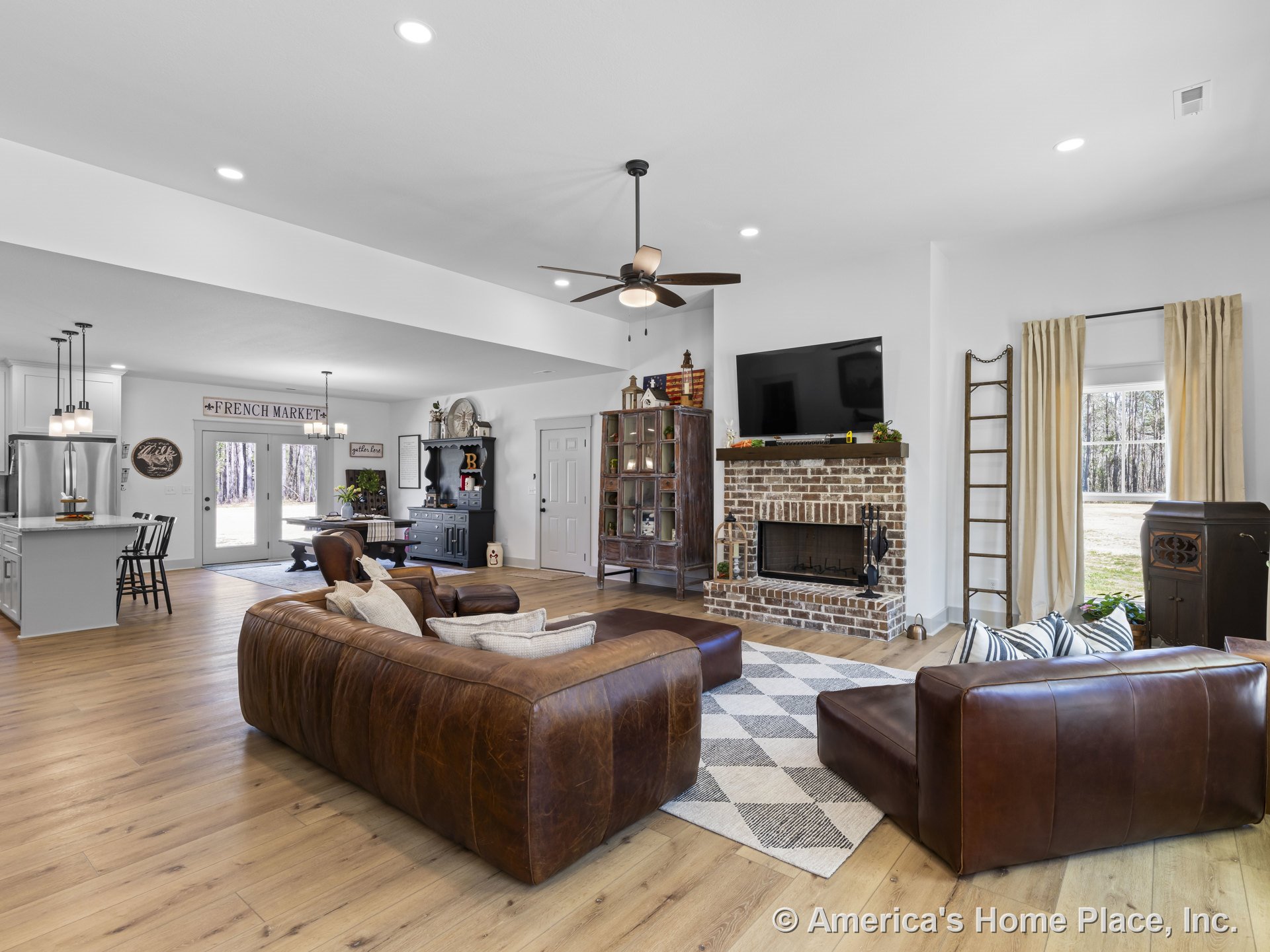 Brick fireplace with wood mantel, light oak plank flooring, large windows with cream curtains, recessed ceiling lights, open kitchen with island, double glass doors leading to