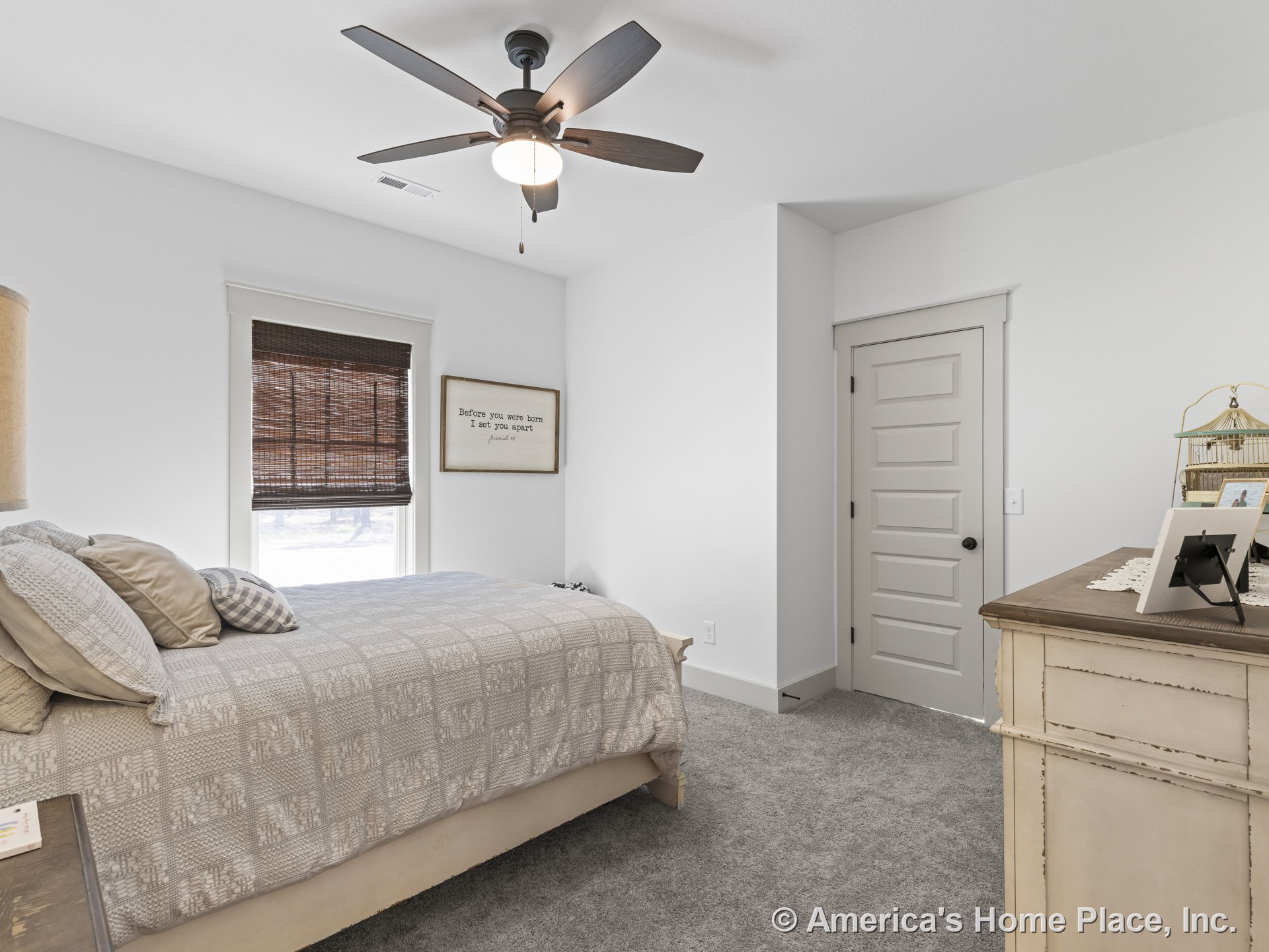 Carpeted bedroom with white walls, paneled interior door, white baseboards and trim, ceiling fan with built-in light, trimmed window with woven shade, and closet door.