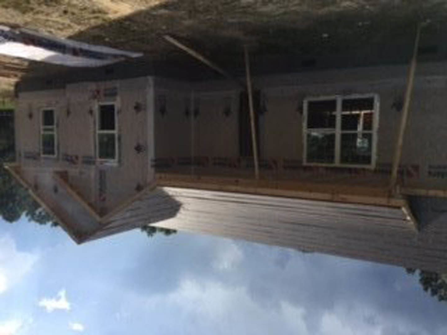 Framed house under construction beside a calm lake, reflected in water; unfinished wood exterior, square-paned windows, cloudy sky overhead