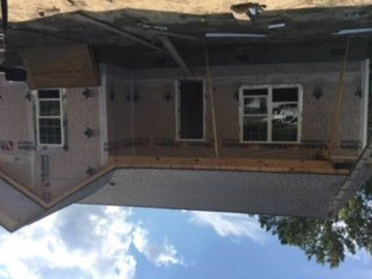 Modern home exterior with large window framed in white trim, reflecting sky and trees, surrounded by light-colored siding and landscaped greenery.
