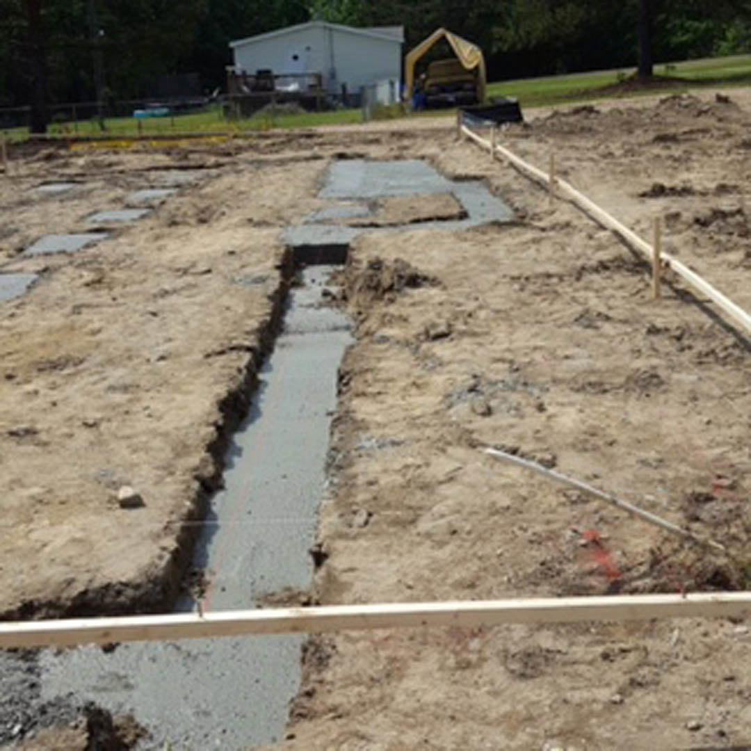 Construction site with exposed soil trench beside partially built house, surrounded by grass, trees, and scattered building materials.