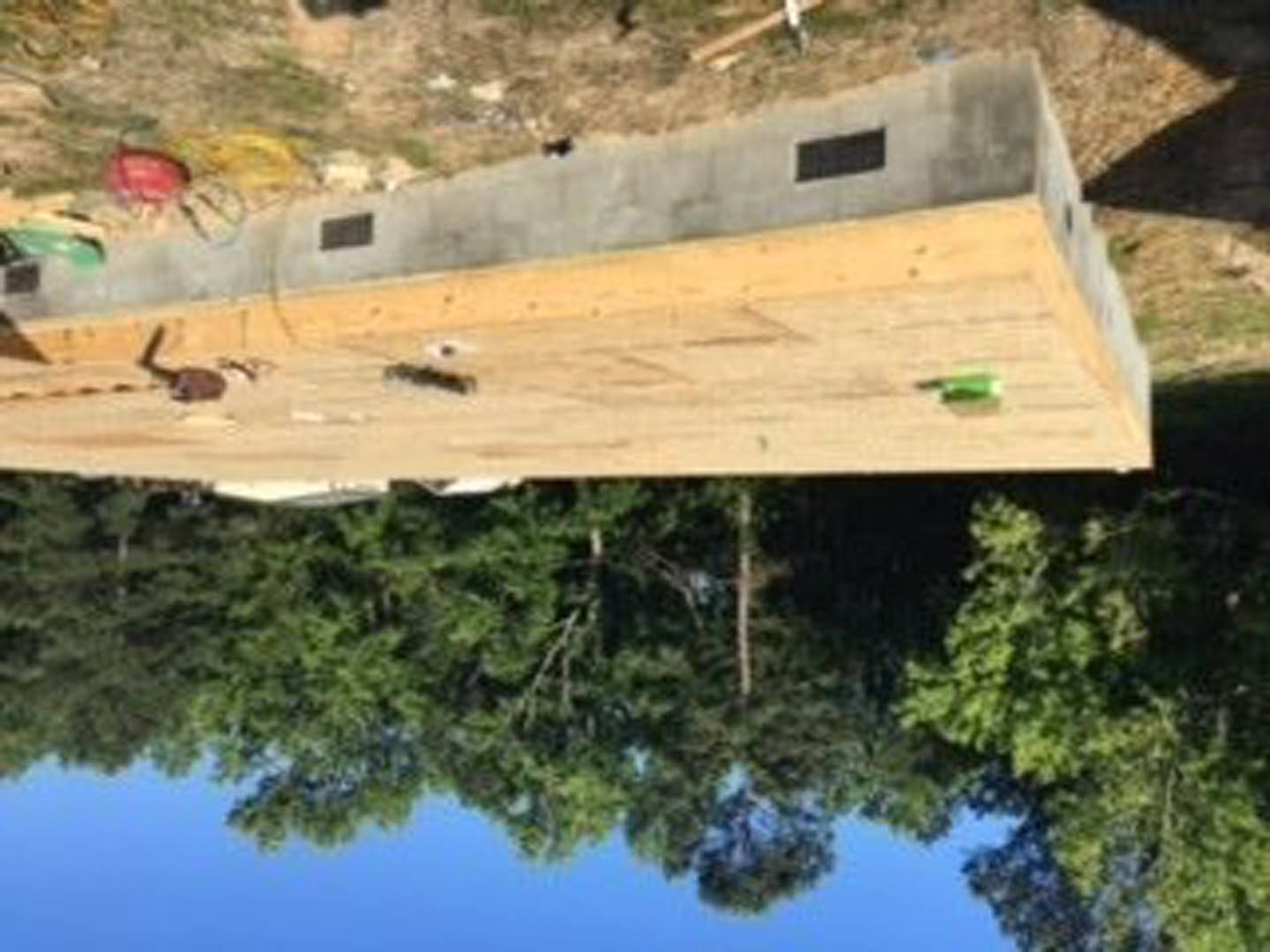 Concrete foundation walls with circular cutouts, wooden framing in progress, surrounded by mature trees under a clear blue sky