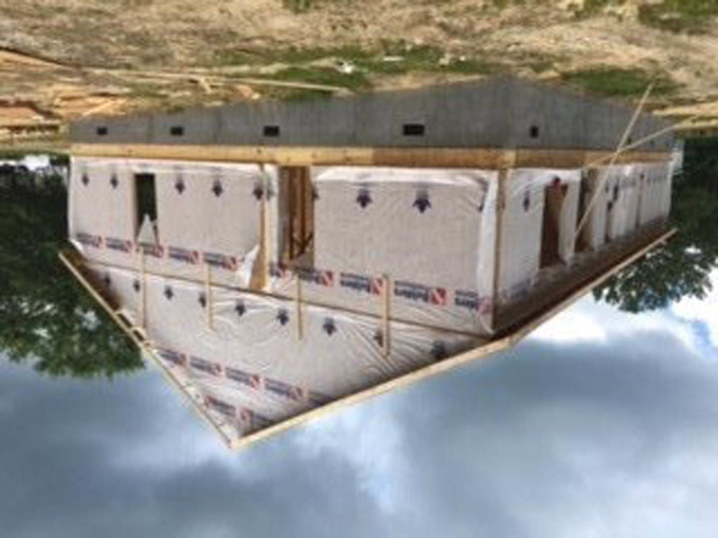 Modern house under construction with triangular roof, white exterior walls, black and red signage, large windows reflecting clouds, surrounded by trees and open sky