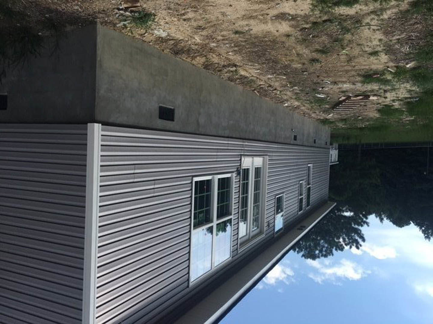 Modern home with light stucco walls, large windows reflecting sky, double entry doors, square wall vent, and backyard pool overlooking a dirt and grass field near a lake.