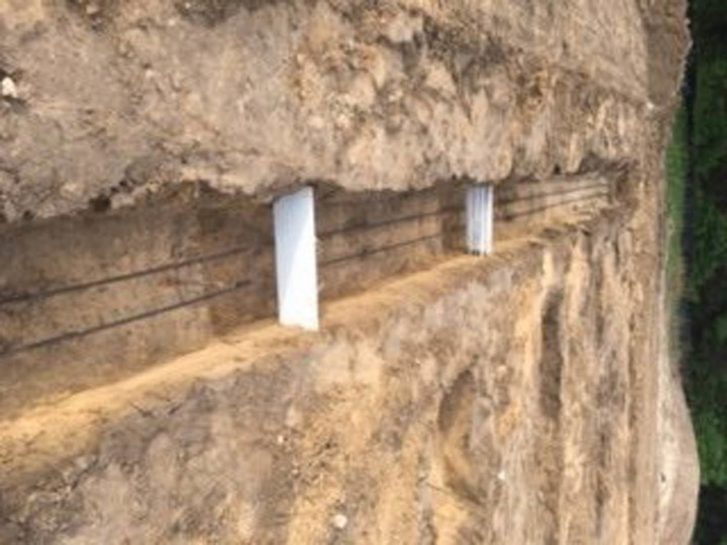 Rough limestone wall with visible metal rods and bars, white pole featuring a black stripe, rocky ground in foreground, outdoor setting with blurred building and mountain in