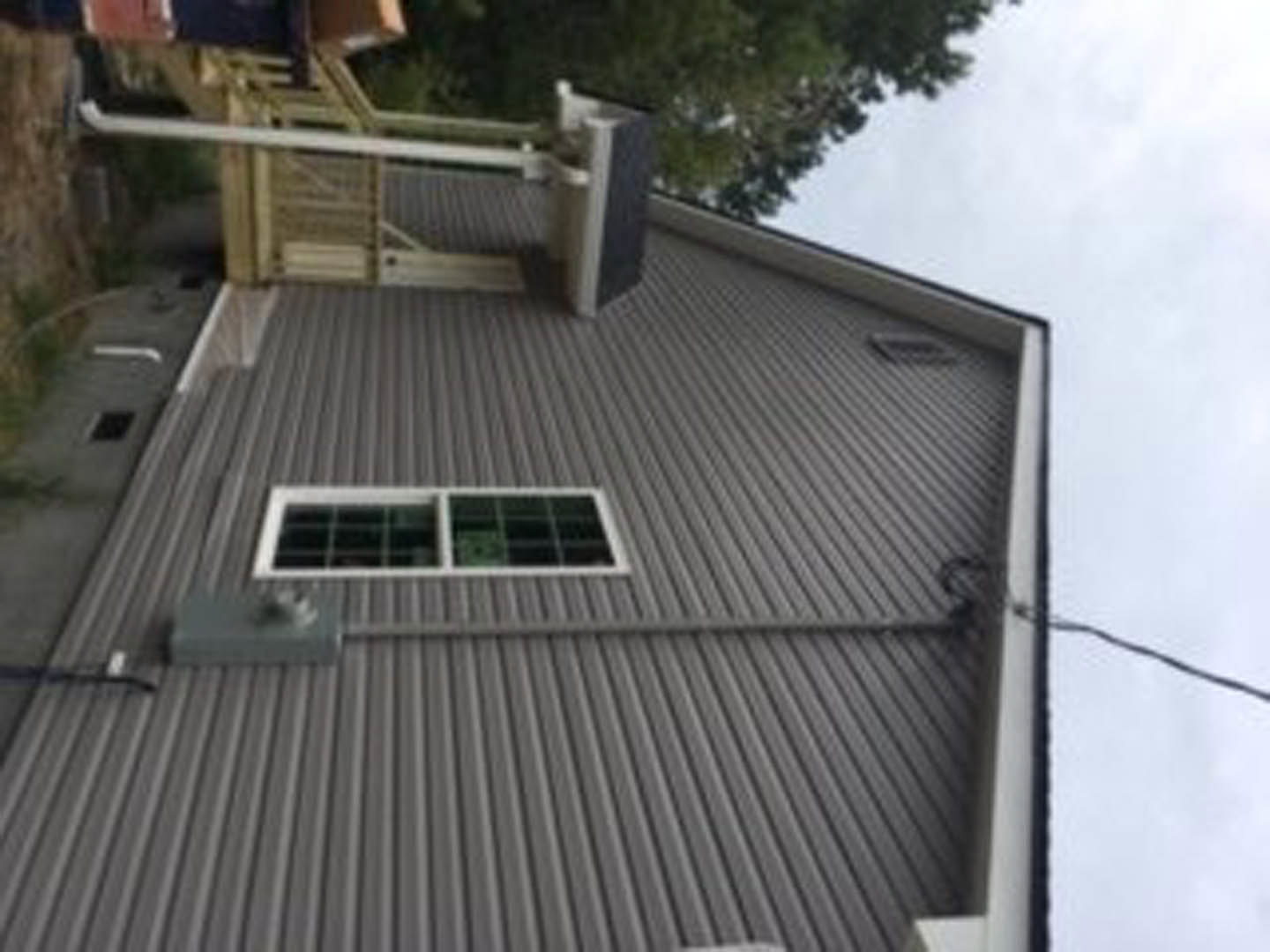 Grey siding with white metal roof, large window, deck railing, tree branches, partial view of a person’s leg