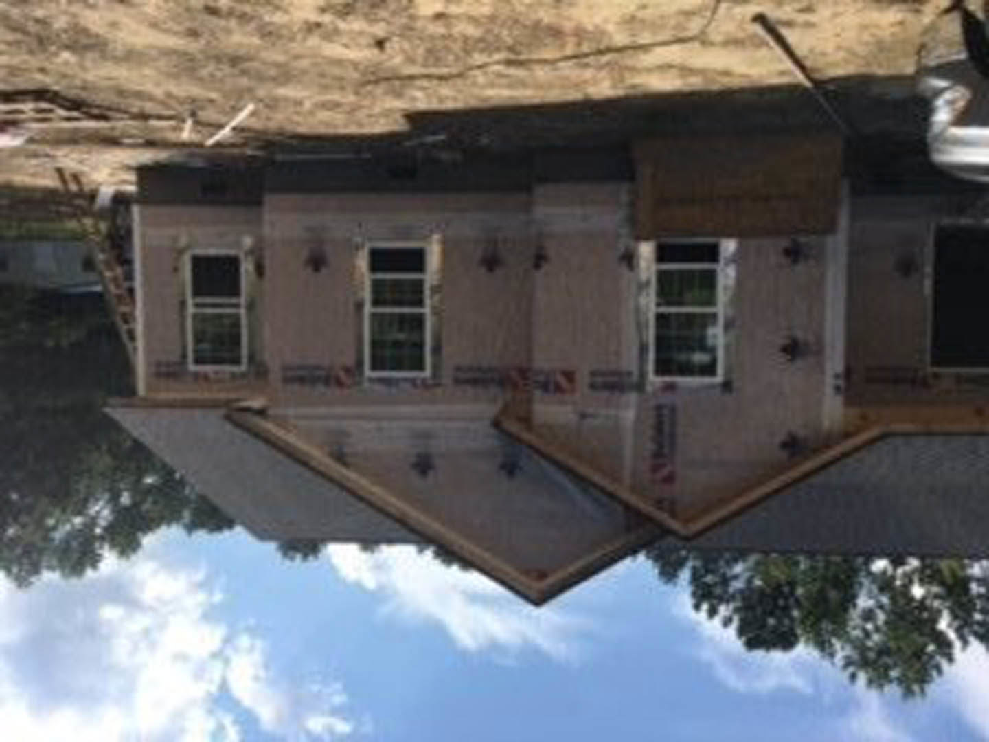 Partially built house with exposed wooden framing, white-framed windows, and roof sheathing, surrounded by tall trees under a cloudy sky