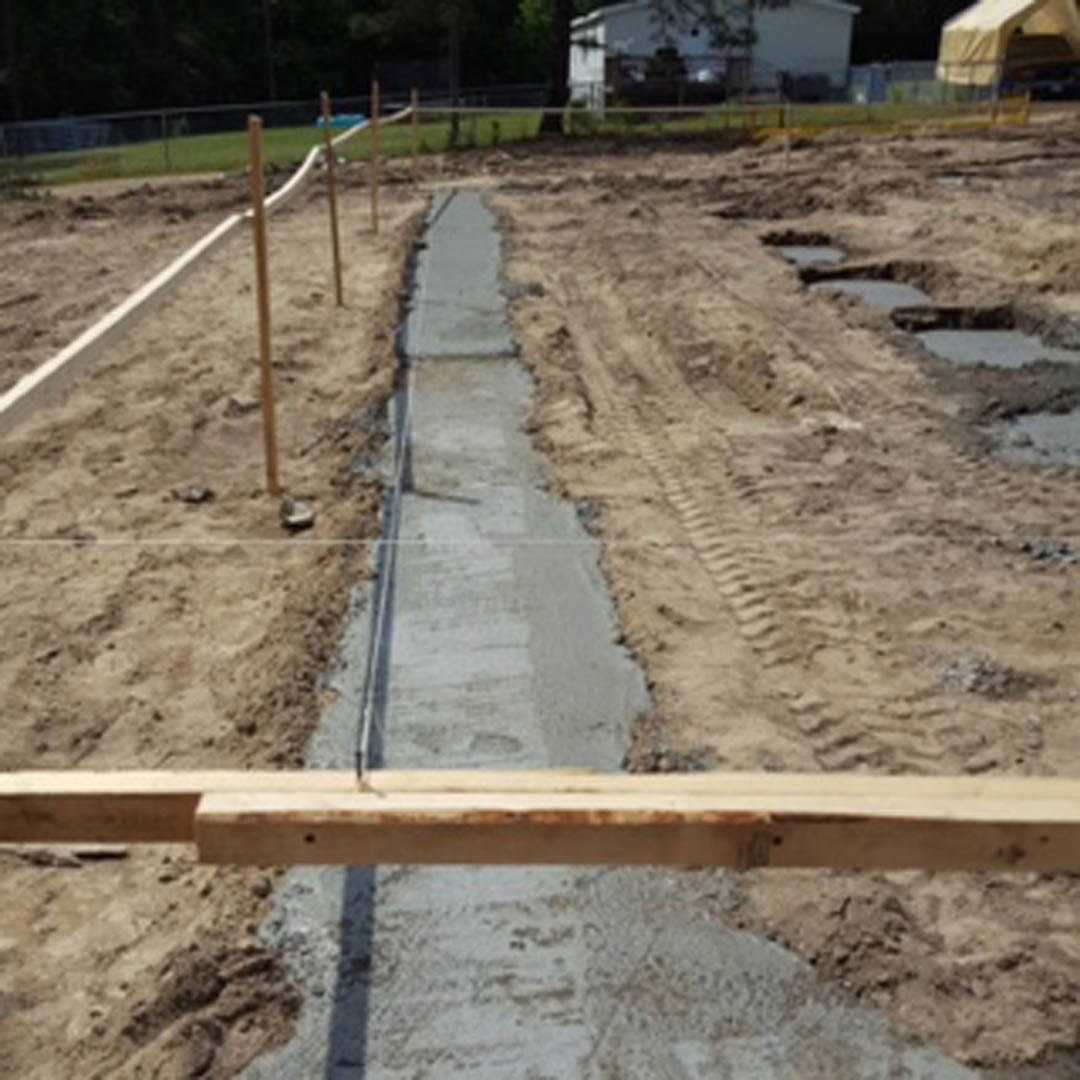 Construction site with exposed soil foundation, wooden fence, scattered building materials, and trees in the background