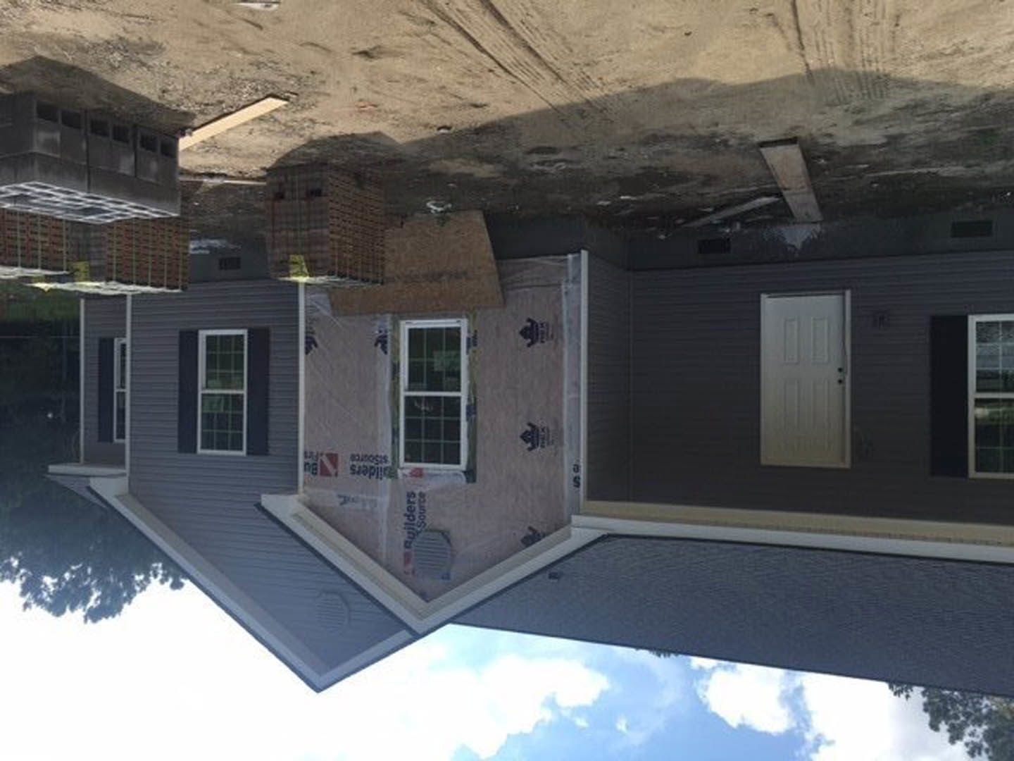 Partially finished exterior of a house with white-framed windows, white door featuring a black handle, scattered cardboard boxes near the entrance, and a black logo on a white