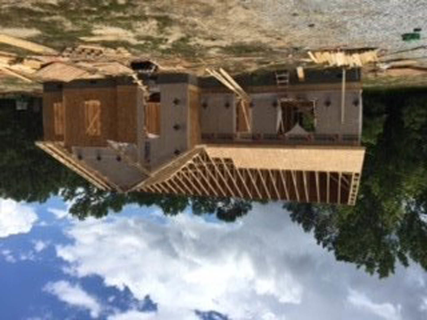 Wood-framed house under construction with exposed beams, surrounded by trees under a clear blue sky