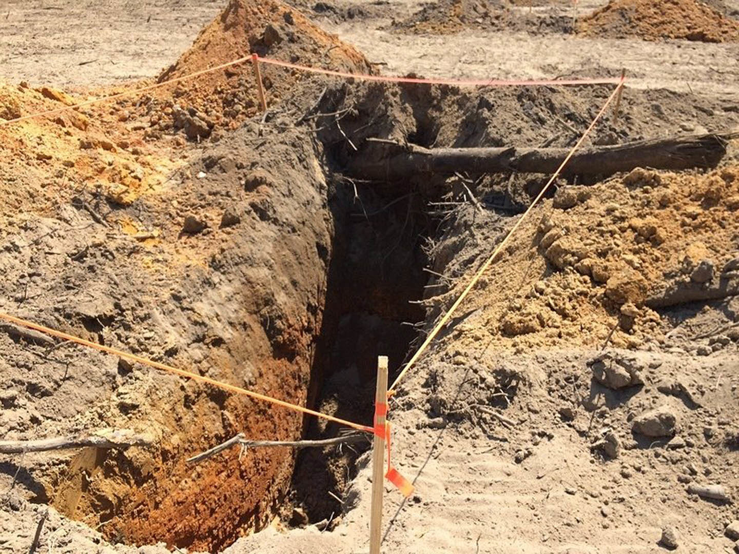 Freshly excavated hole in compacted soil with exposed rocks and a rope partially buried in dirt, outdoor residential construction site