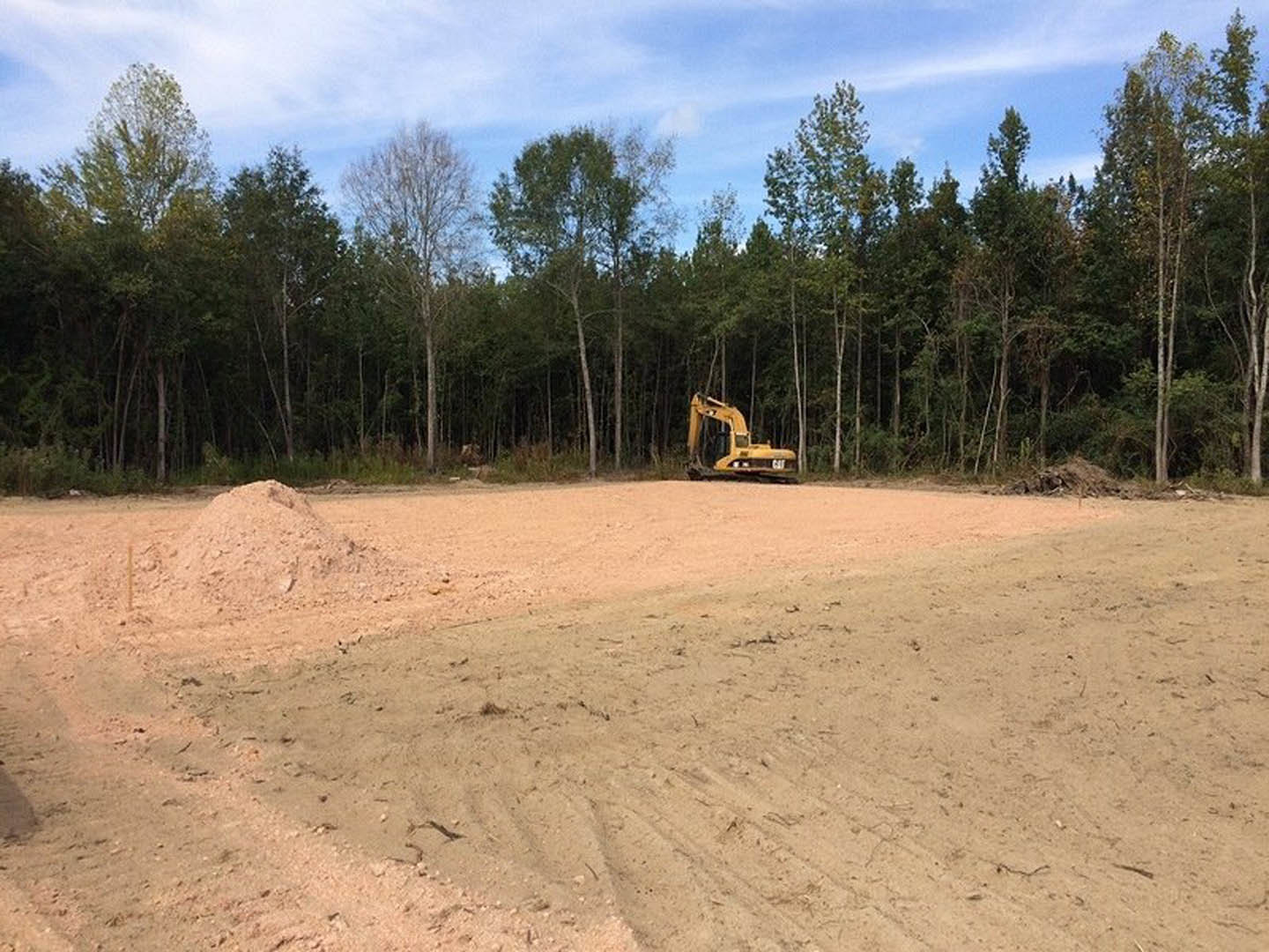 Yellow excavator parked on dirt mound at residential construction site, surrounded by bare trees and blue sky