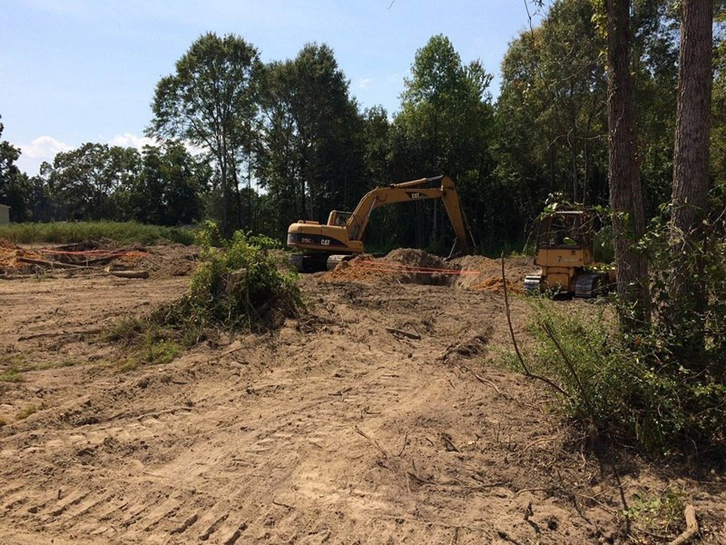 Bulldozer and tractor on dirt construction site surrounded by trees and bushes, exposed soil and tree stumps visible