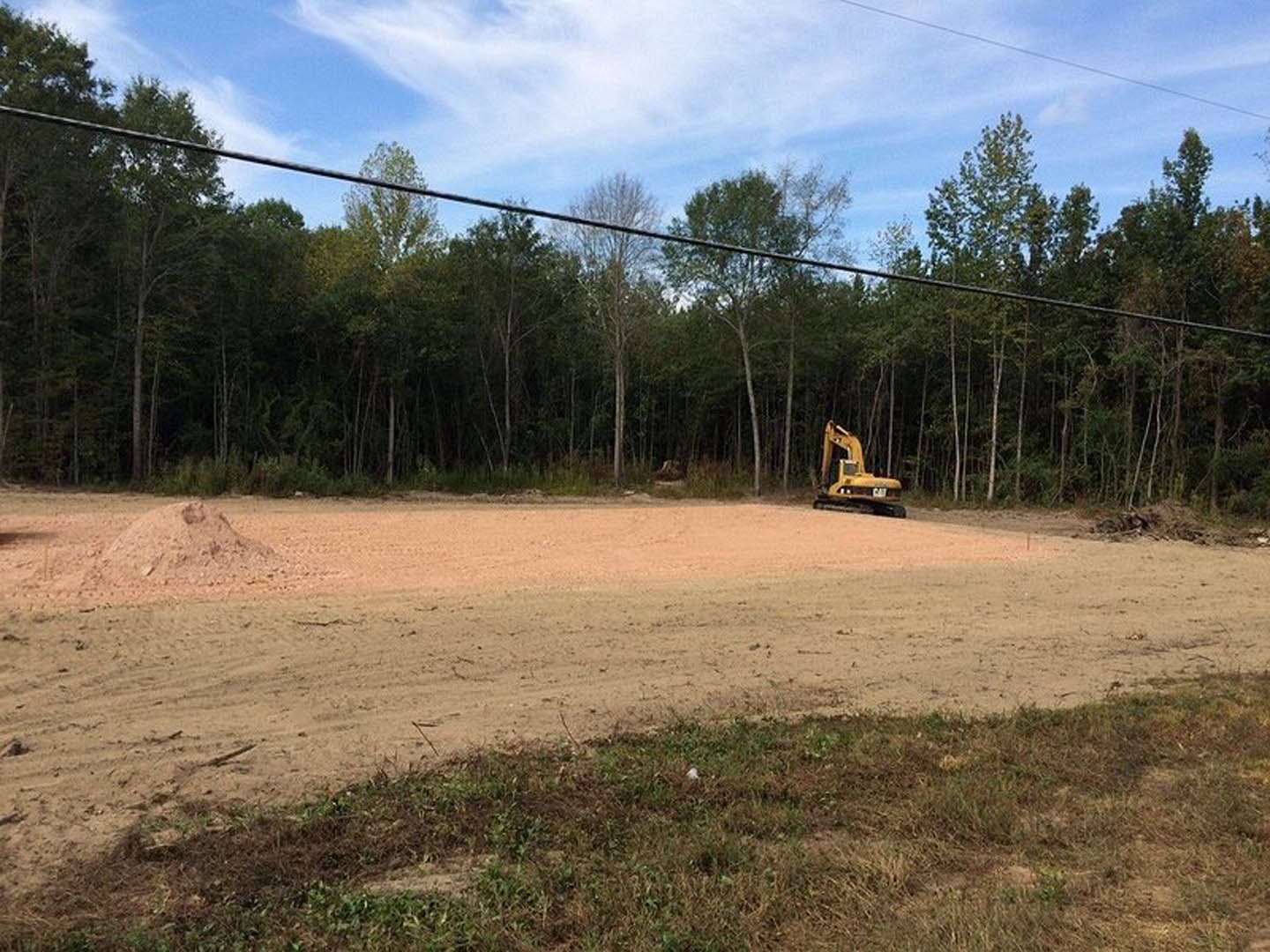 Yellow excavator parked on dirt field surrounded by grass, trees, and blue sky with scattered clouds