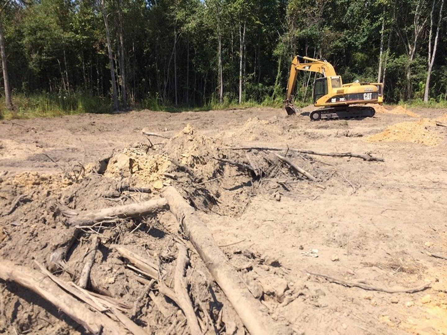 Yellow bulldozer parked on dirt lot beside piles of soil and logs, surrounded by trees and forested landscape