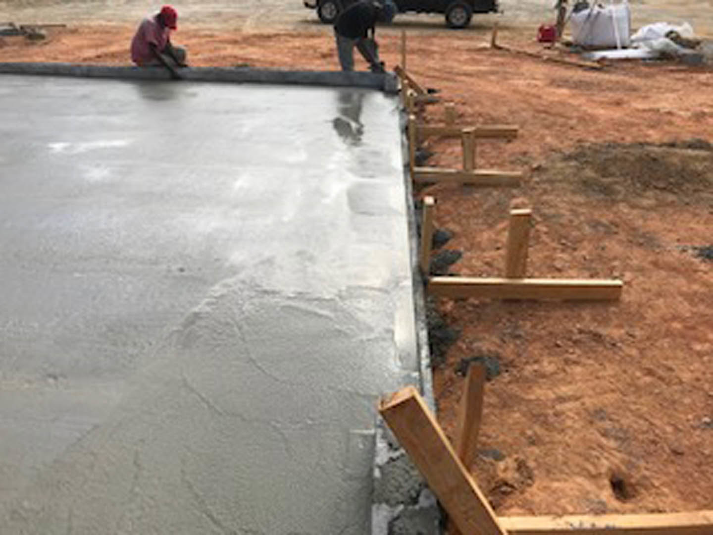 Workers installing concrete slab foundation at residential construction site, surrounded by dirt and wooden framing, with tools and materials scattered outdoors.