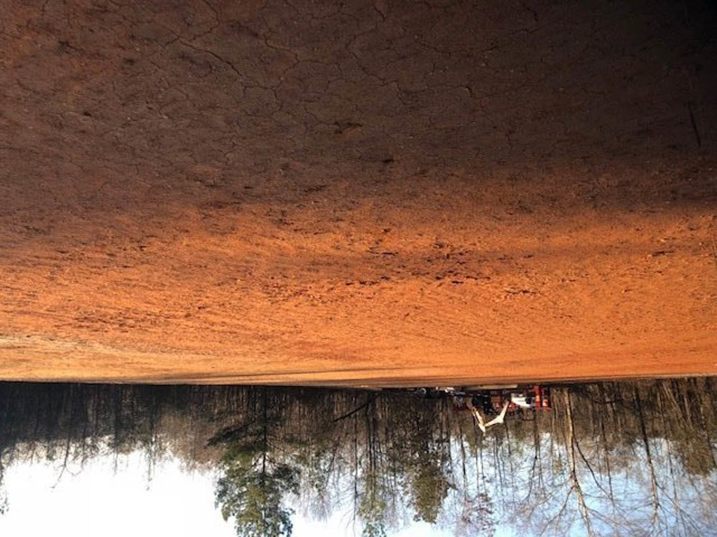 Dirt road lined with leafy trees, tractor parked on dry, cracked ground, lake reflecting surrounding landscape