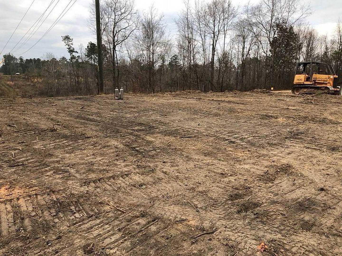 Dirt field with tire tracks, yellow bulldozer parked near edge, group of trees in background, scattered grass and plants along soil