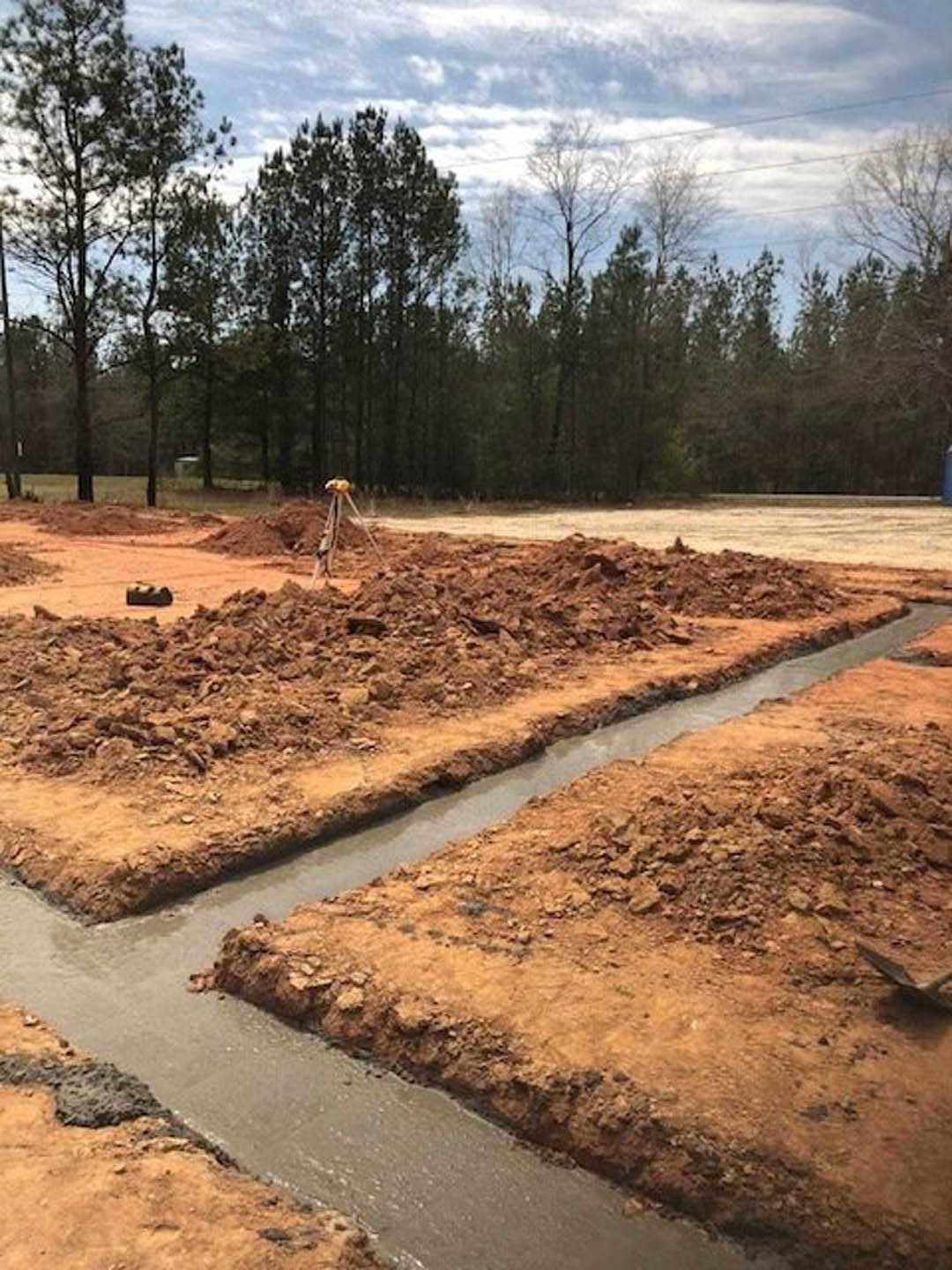 Dirt construction site bordered by mature trees, exposed soil and trench visible under cloudy sky