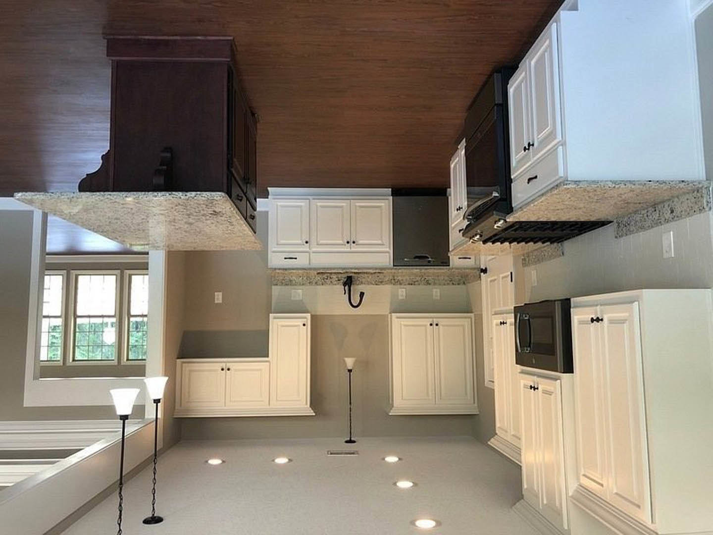 White kitchen cabinets, black microwave above white countertop, stainless steel sink, white walls, window, white door with black handle, lamp on pole, black chain hanging from long