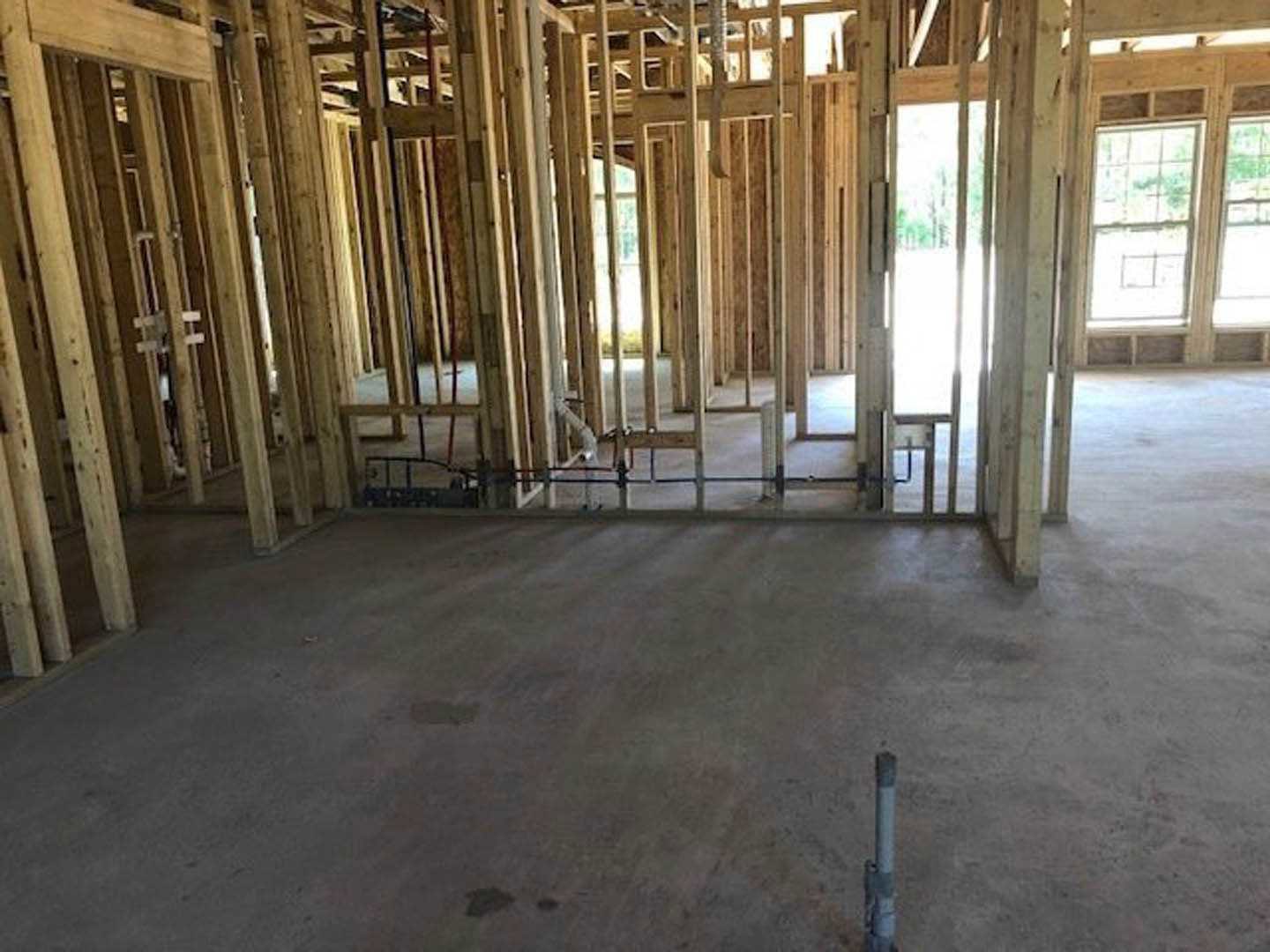 Wood-framed room under construction with exposed beams, concrete floor featuring visible plumbing pipes, multi-pane window, and close-up details of framing and support pole.
