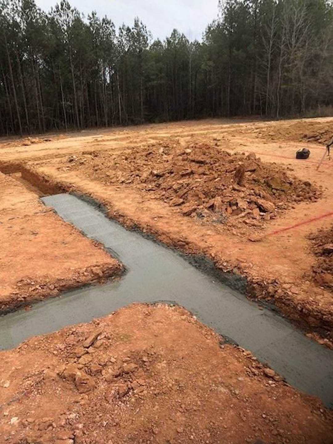 Dirt trench bordered by soil piles, surrounded by tall trees under a clear sky