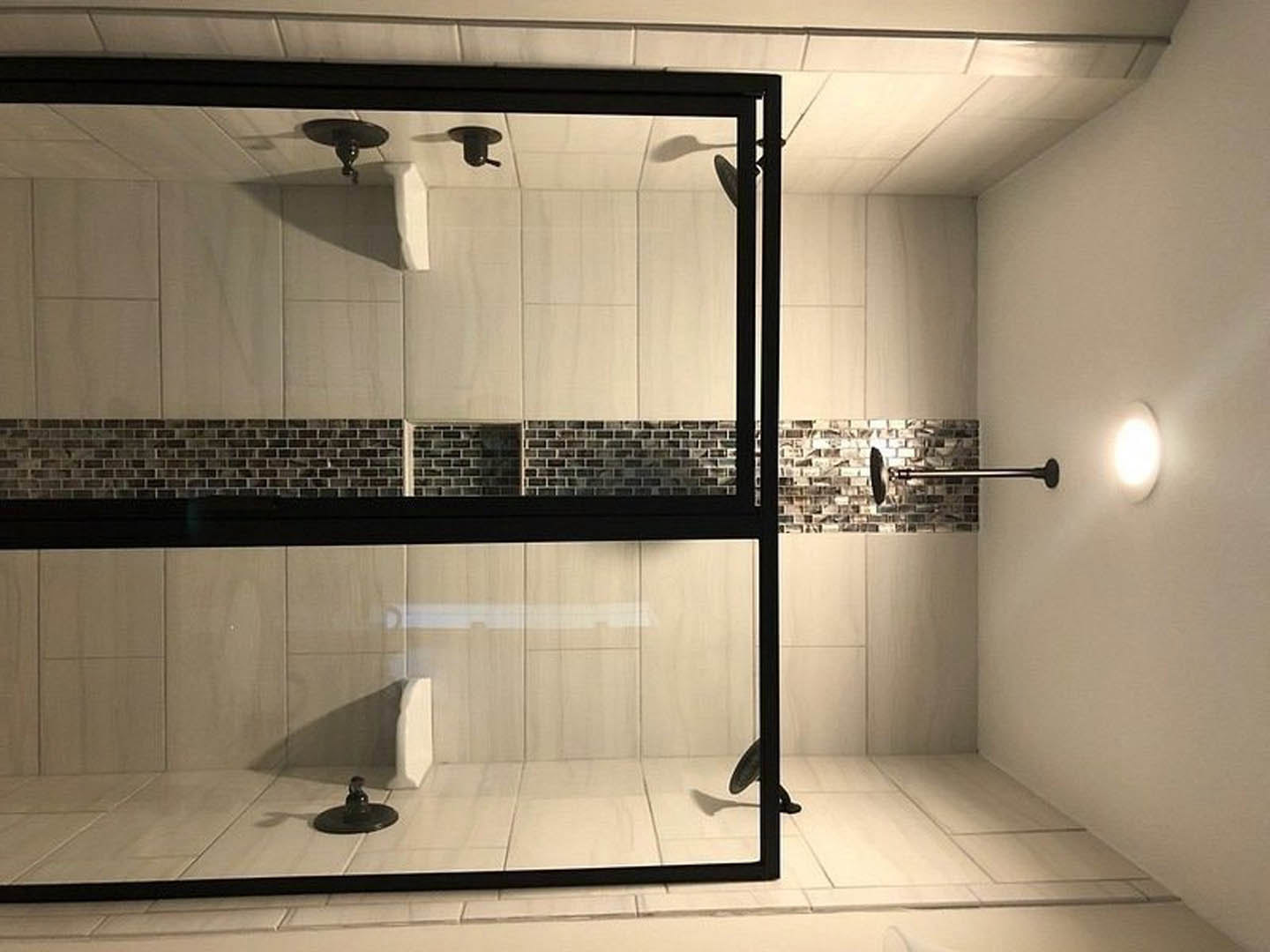 Walk-in shower with clear glass door, large wall tiles, chrome fixtures, and bright overhead lighting; black round side table and decorative bird shadow on tile floor.