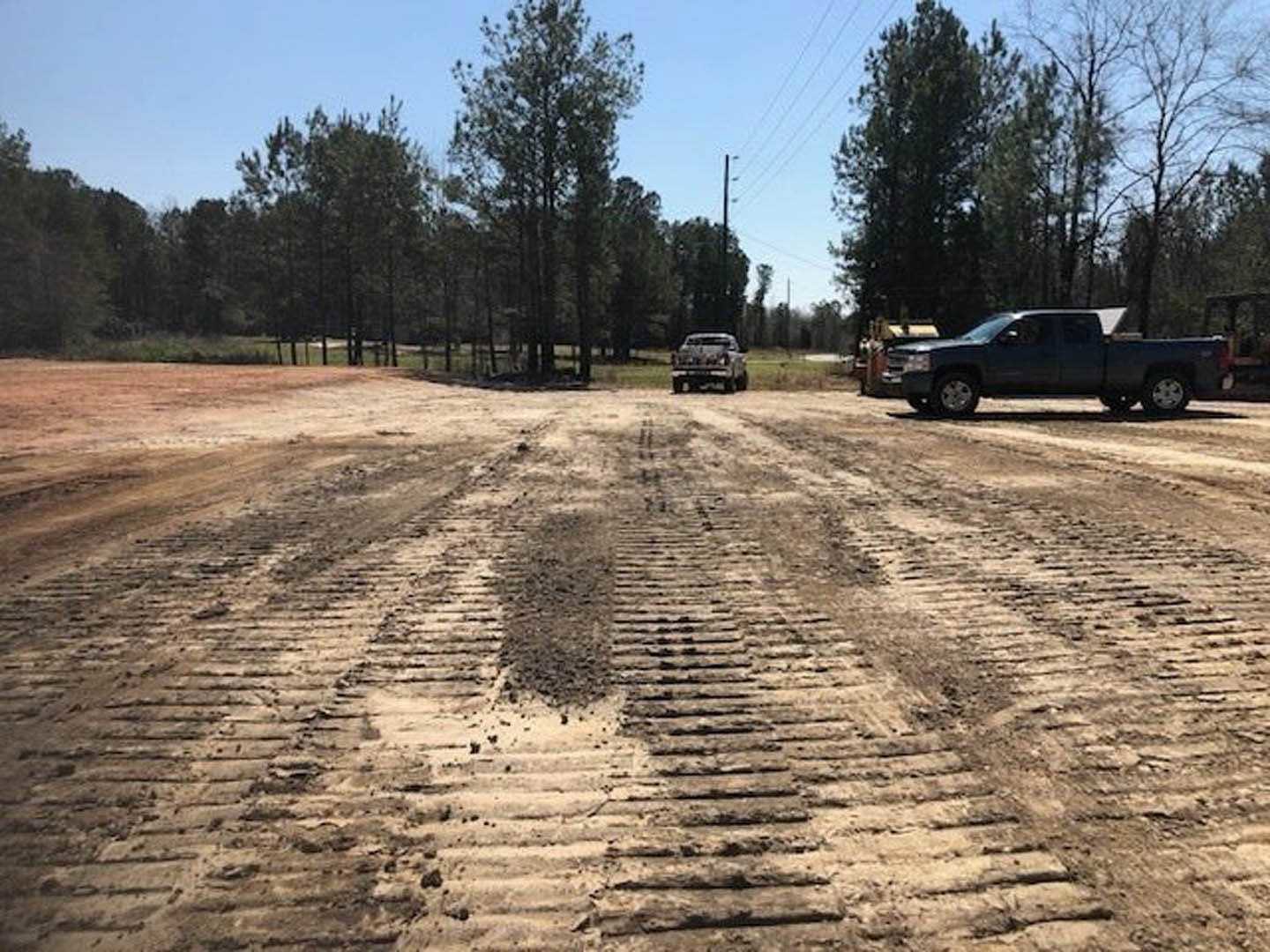Black pickup truck parked on dirt lot beside unfinished ground, tire tracks visible in soil, trees and open sky in background