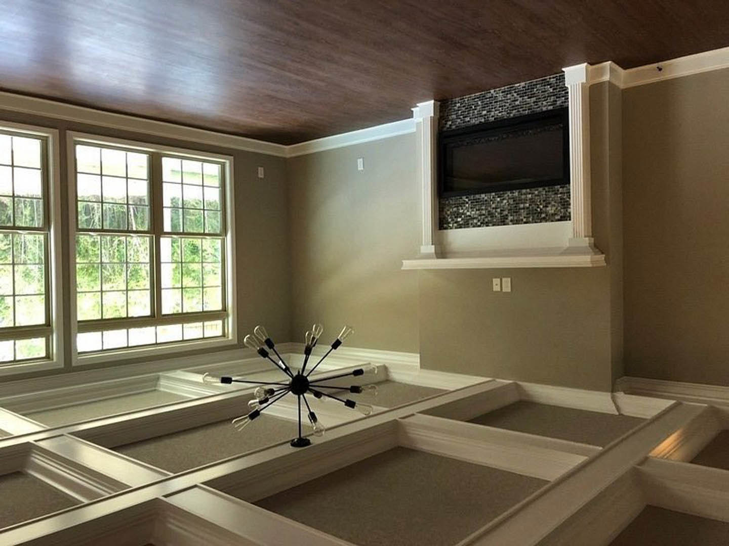Living room with exposed brick wall, mounted flat-screen TV, modern spoke-style chandelier, large window, and neutral finishes.