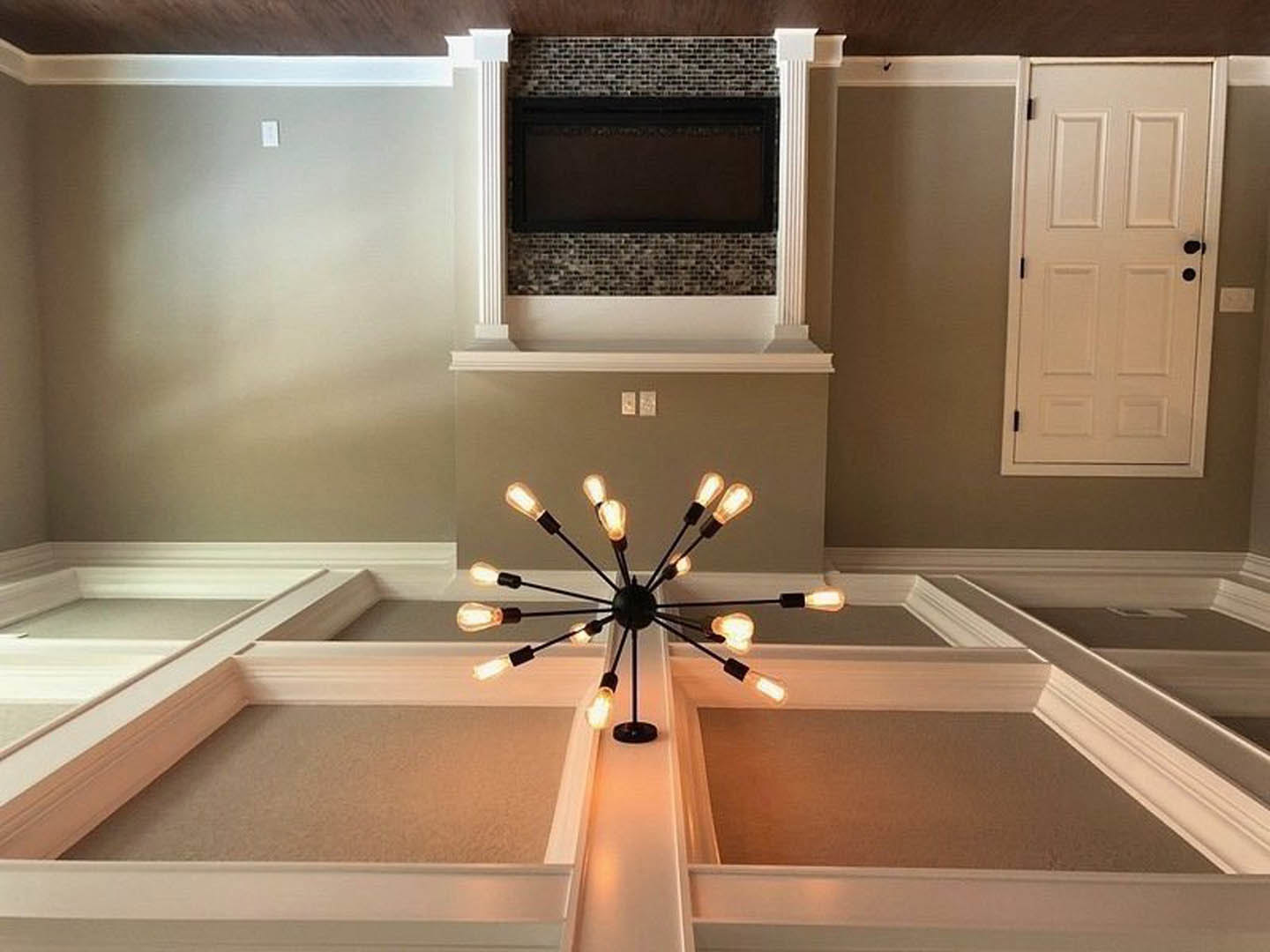 Open kitchen with white cabinetry, marble countertops, stainless steel sink and tap, brick accent wall with mounted TV, chandelier hanging from ceiling, white door with black